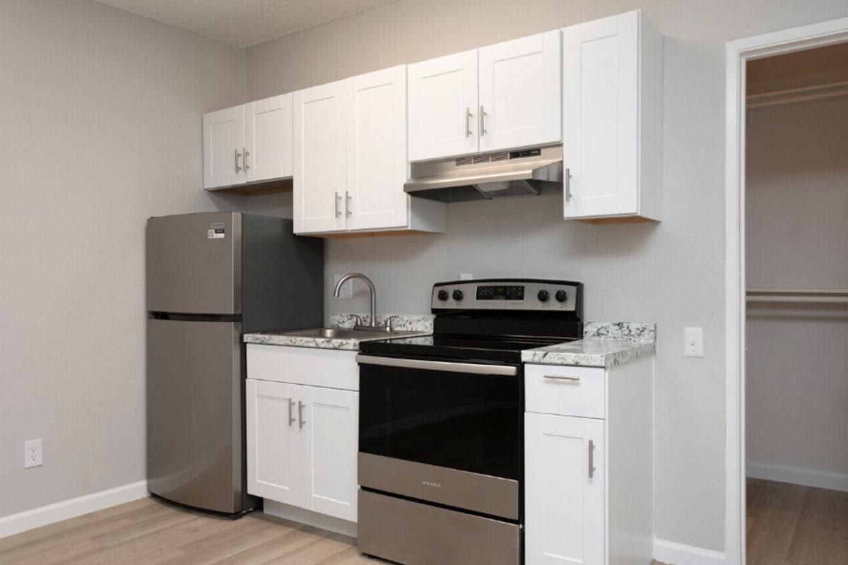 A modern kitchen featuring white cabinets, a stainless steel refrigerator, an electric stove with an oven, and a stainless steel sink. The countertops are made of granite. The walls are painted in a light grey color, and there is a door leading to a closet in the background.