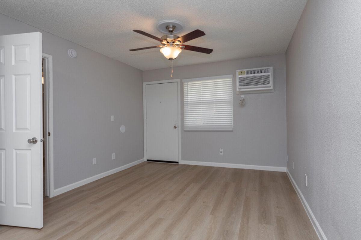 A small, empty room with light gray walls and wooden flooring. It features a ceiling fan, an air conditioning unit on the wall, and a window with blinds. A closed door leads to an adjacent space, and the room is well-lit with natural light.