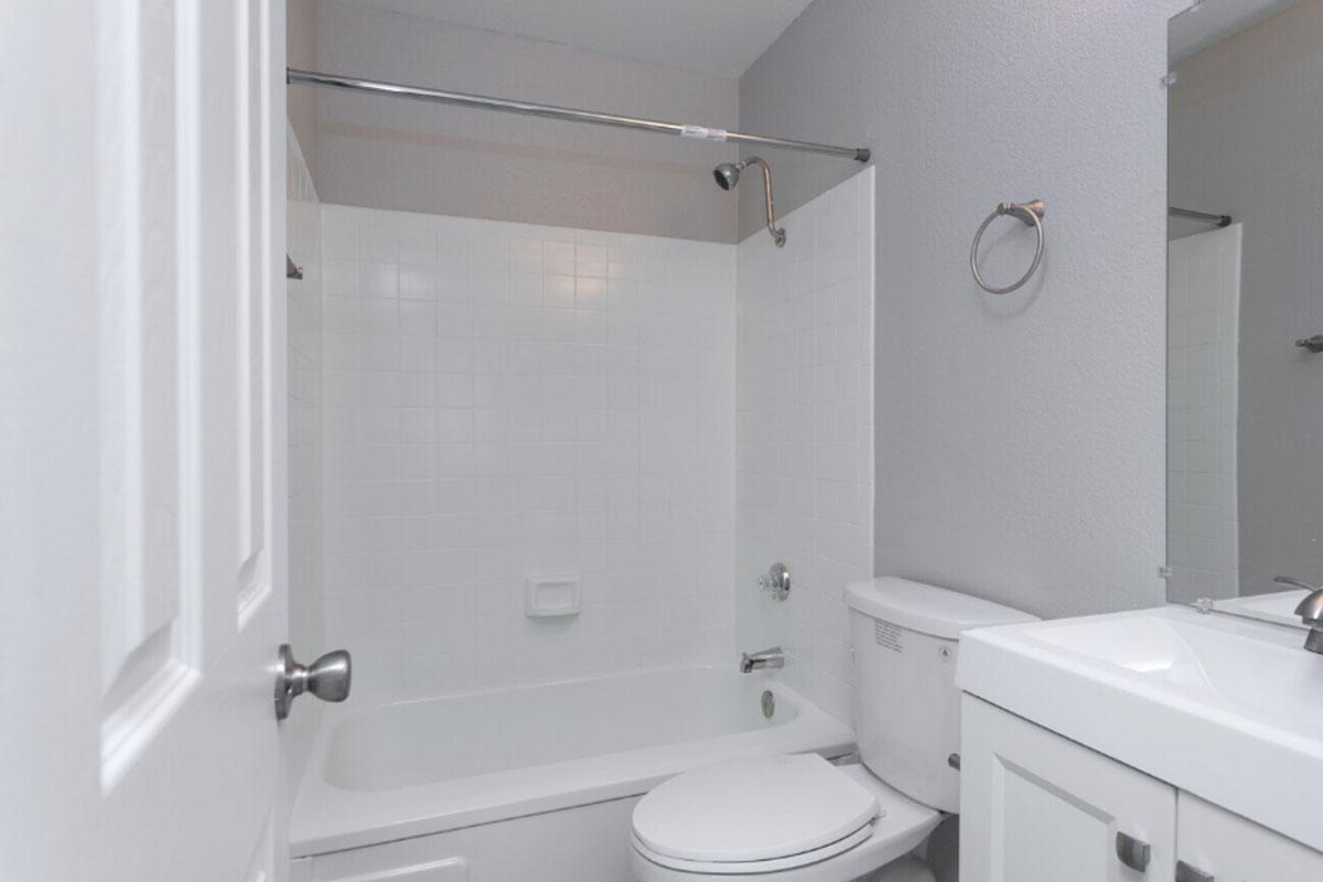 A clean and modern bathroom featuring a white bathtub with a shower curtain, a toilet, and a sink with a mirror above it. The walls are painted in a light grey color, and the floor has a neutral tone, creating a bright and fresh atmosphere.