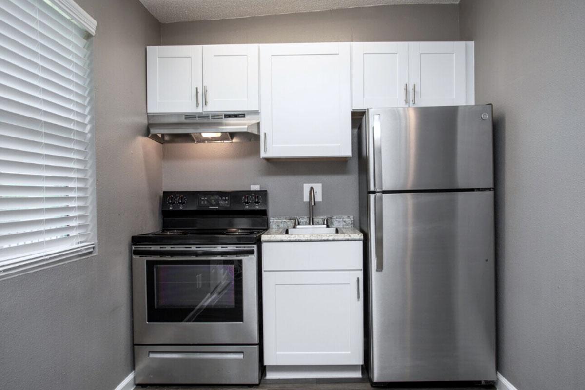 Modern kitchen with gray walls, white cabinets, a stainless steel refrigerator, a black oven with a smooth top, and a white countertop. The window has blinds allowing natural light. The space is clean and minimalist, ideal for a small apartment or home setting.