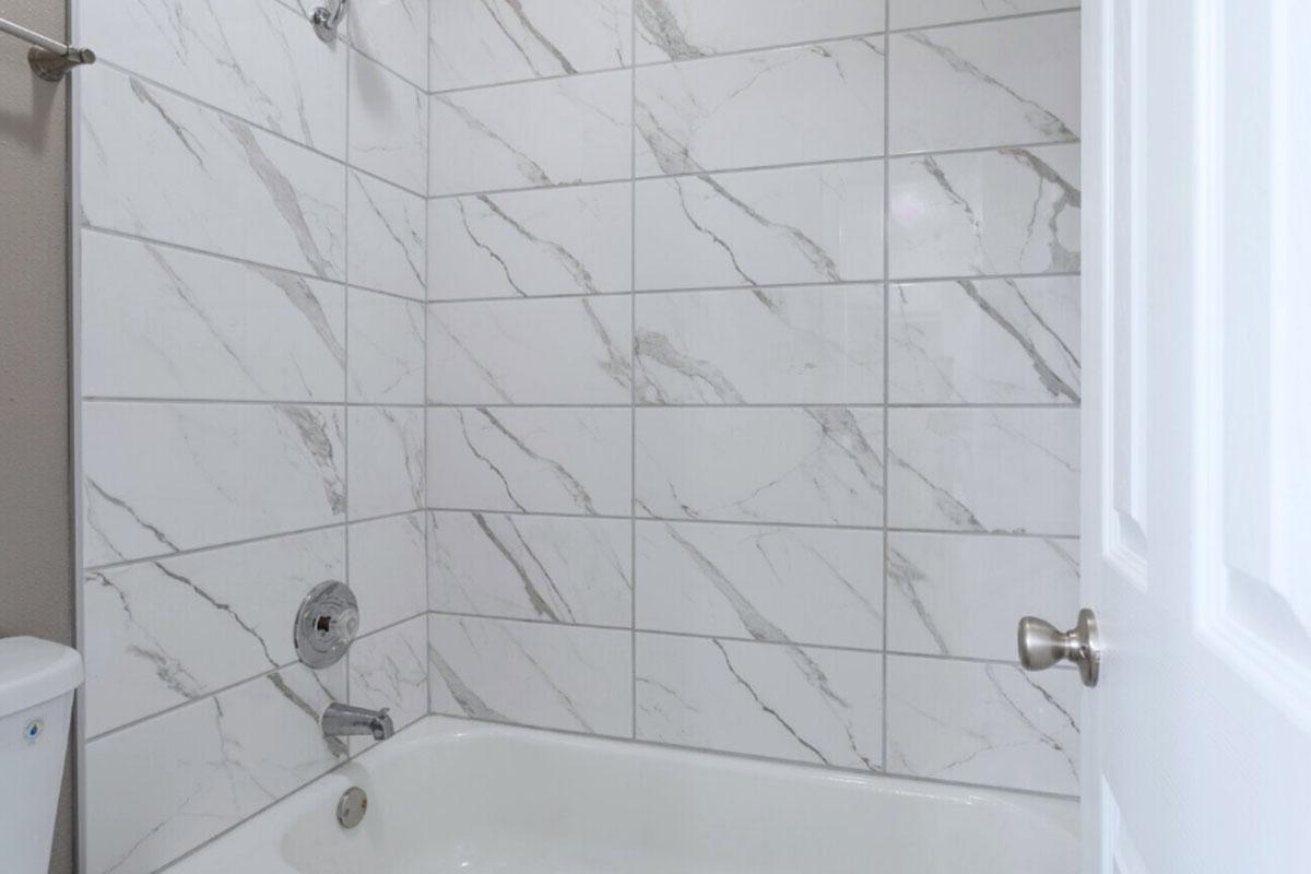 A clean bathroom with a bathtub and a shower. The walls are covered in large, white marble-patterned tiles. A chrome showerhead is mounted on the wall, and there is a faucet for the bathtub. A white door is partially visible on the right side. The overall appearance is modern and tidy.