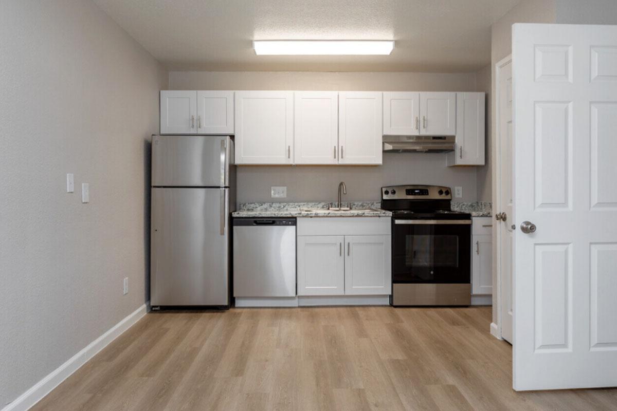 Modern kitchen with white cabinets, stainless steel appliances including a refrigerator, dishwasher, and stove. Countertops are speckled with gray and white. The floor is light wood laminate, and the room is well-lit with an overhead light. A door is partially visible on the right.