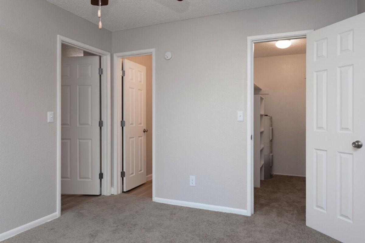 A well-lit room with beige carpet featuring three doors. Two doors lead to adjacent rooms, and one door opens to a small closet. The walls are painted a light gray, and a ceiling fan can be seen at the top of the image. The overall atmosphere is clean and modern.