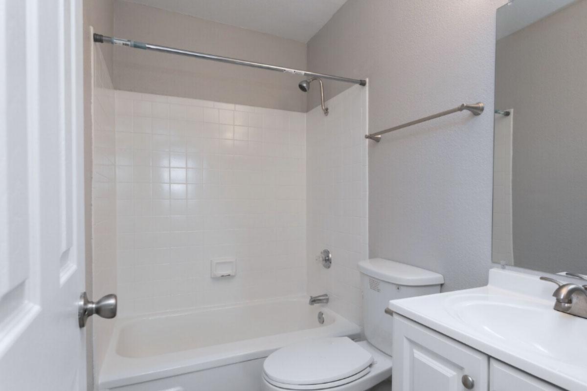A clean and modern bathroom featuring a white bathtub with a sliding shower curtain, a toilet, and a sink cabinet with a mirror above. The walls are painted in a light color, and the room has a bright and airy feel.