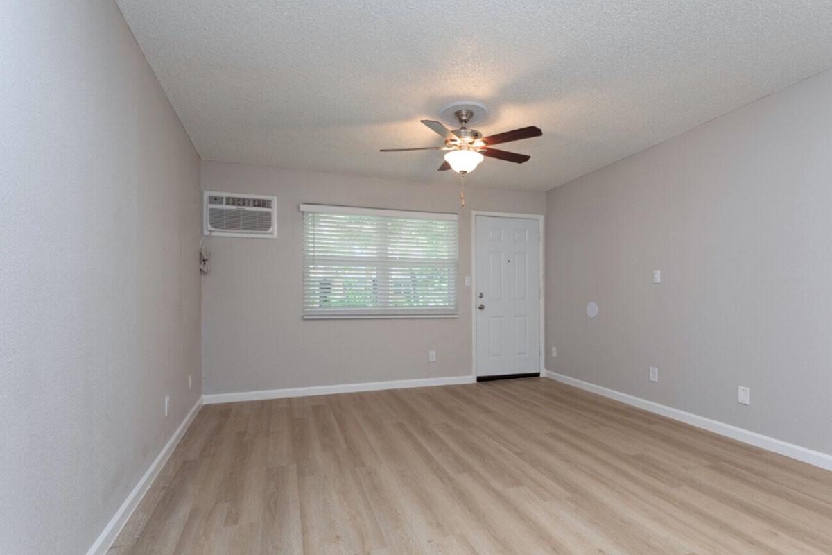 A spacious, empty living room with light-colored walls and a wooden floor. There is a ceiling fan in the center and a window with blinds allowing natural light. An air conditioning unit is mounted on the wall. The room features a front door on one side, creating a welcoming atmosphere.