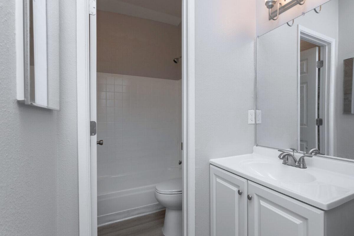 A small bathroom featuring a toilet, a white sink with silver faucets, and a shower/tub combo. The walls are painted a light color, and there is a large mirror above the sink. A door leads to a separate area, creating a sense of privacy. Natural lighting is provided by a wall-mounted light fixture.