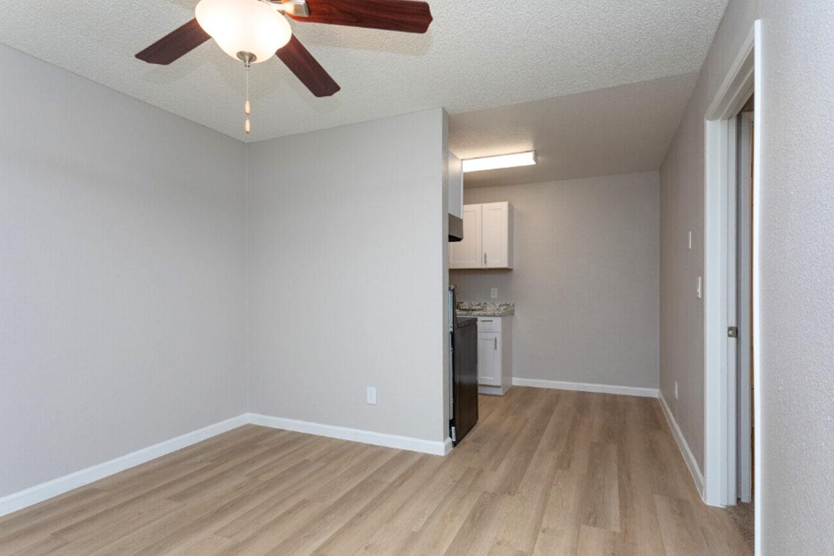 A light-filled living space with a ceiling fan, wooden flooring, and grey walls. In the background, there's a small kitchenette with white cabinets and appliances. The room is spacious and features a doorway leading to another area, enhancing the open layout.