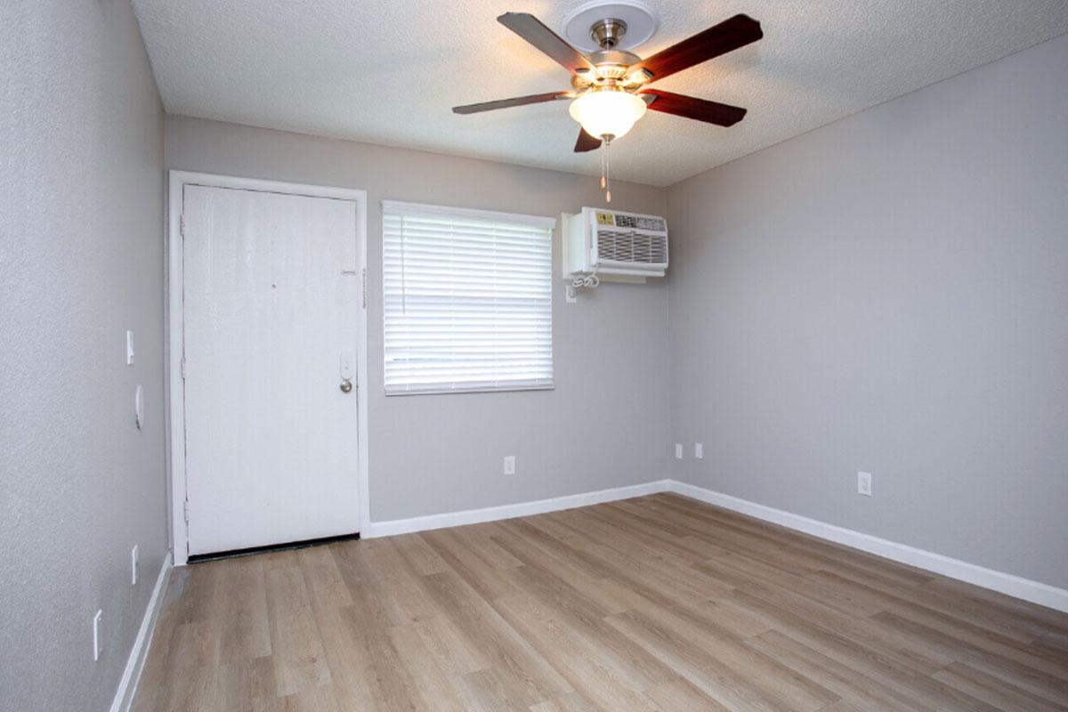 A small, empty room with light gray walls and a ceiling fan. There is a door on one side and a window with blinds opposite it. An air conditioning unit is mounted on the wall near the window. The floor is light-colored wood laminate, creating a bright and open atmosphere.