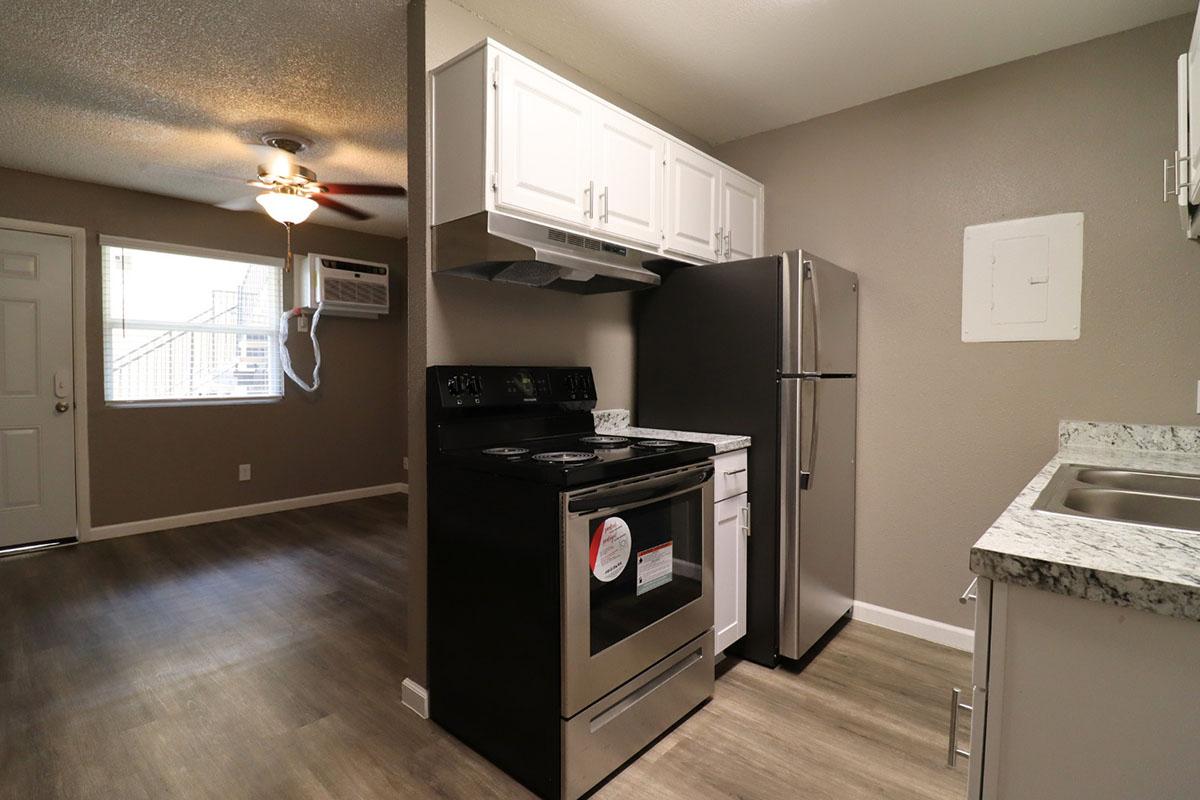 A modern kitchen featuring a stainless steel refrigerator, black stove, and white cabinets. The space has a granite countertop and is connected to a living area with a ceiling fan and an air conditioning unit. Natural light enters through a nearby door leading outside.