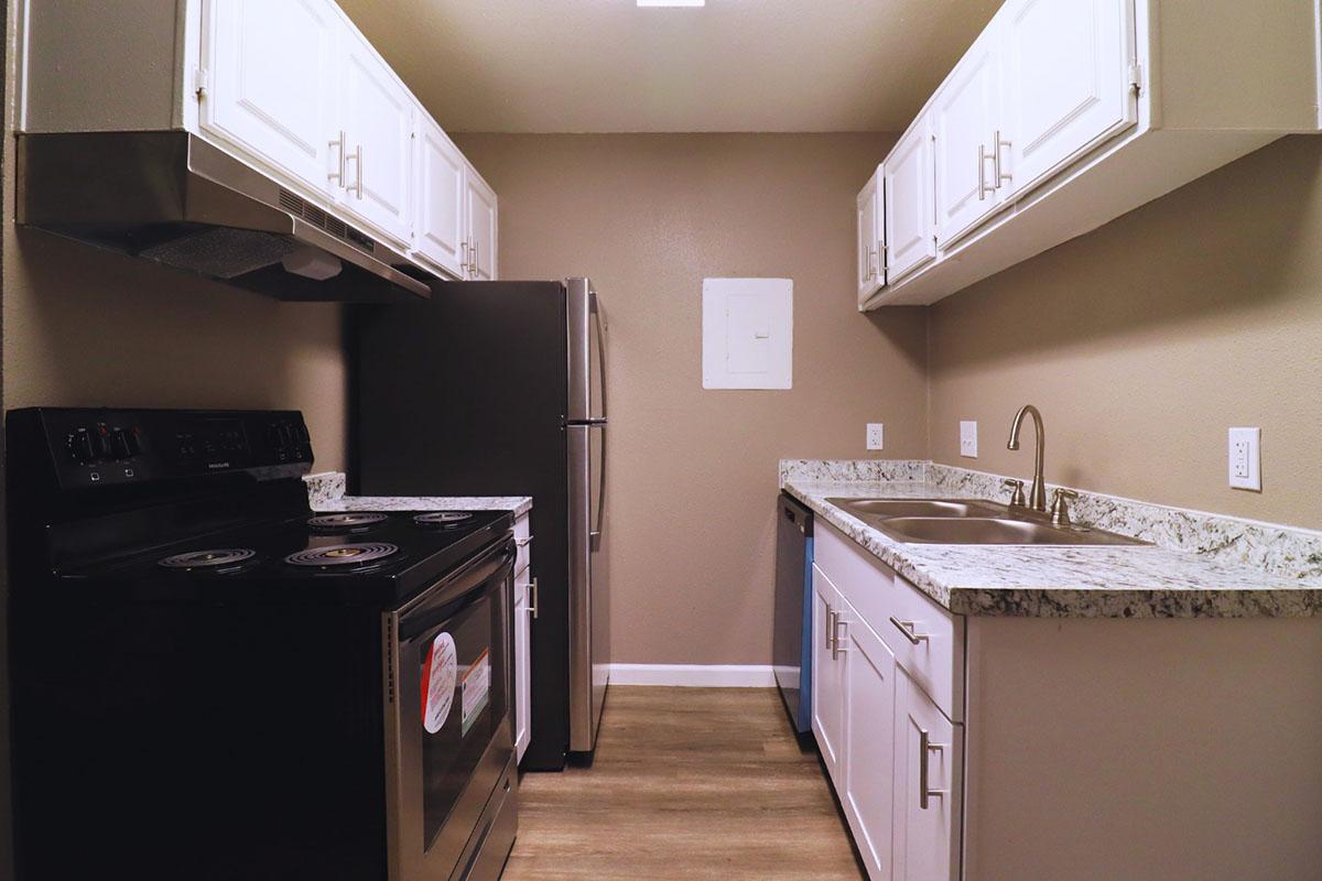 A modern kitchen featuring white cabinetry, stainless steel appliances including a refrigerator and a stove, and a granite countertop. The walls are painted a neutral color, and the floor has a wood-like finish, creating a clean and contemporary look.