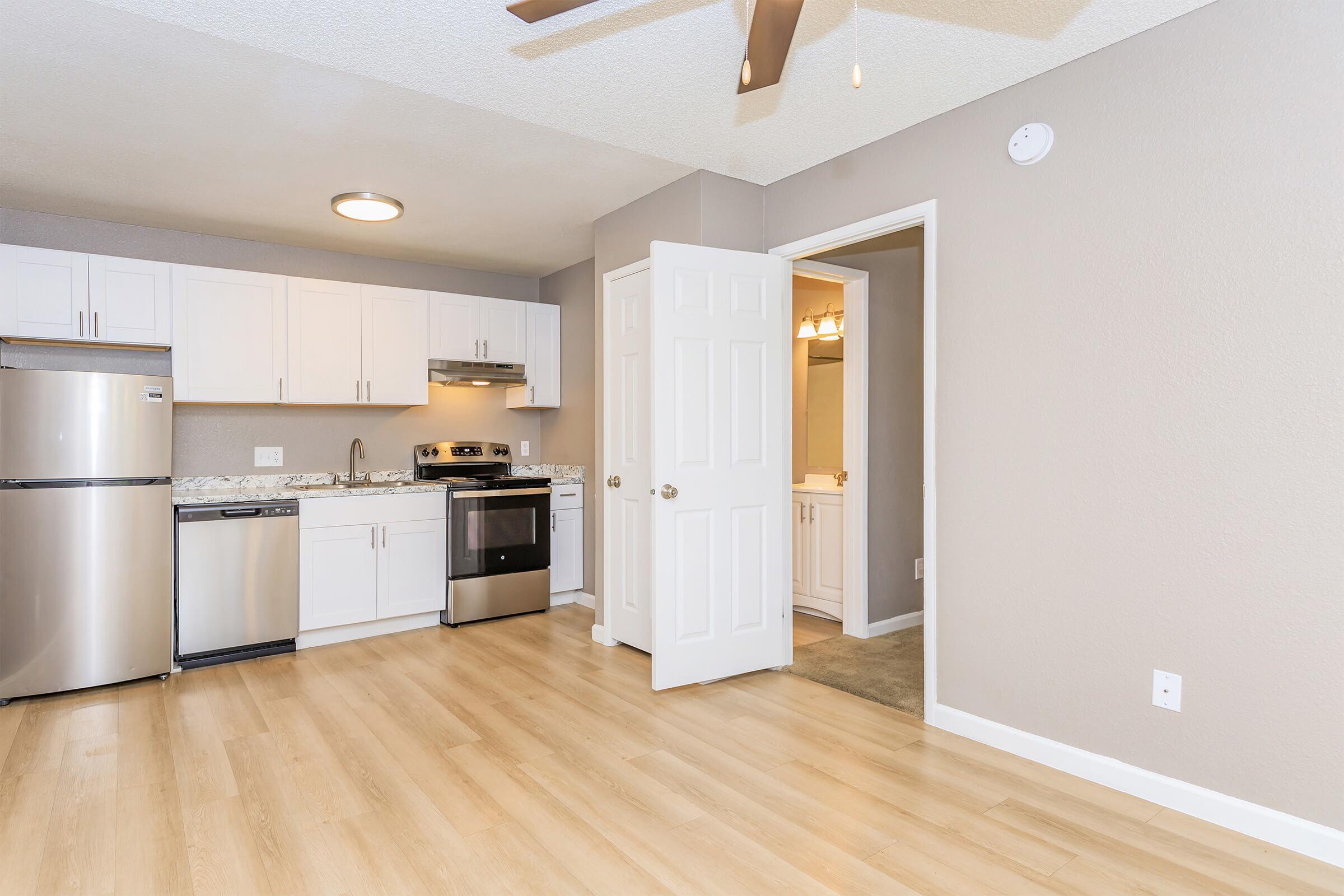 A modern kitchen and living area with light wood flooring. The kitchen features stainless steel appliances, including a refrigerator and dishwasher, along with white cabinets and granite countertops. A doorway leads to a bathroom, and a ceiling fan is visible above. Soft gray walls complete the inviting space.