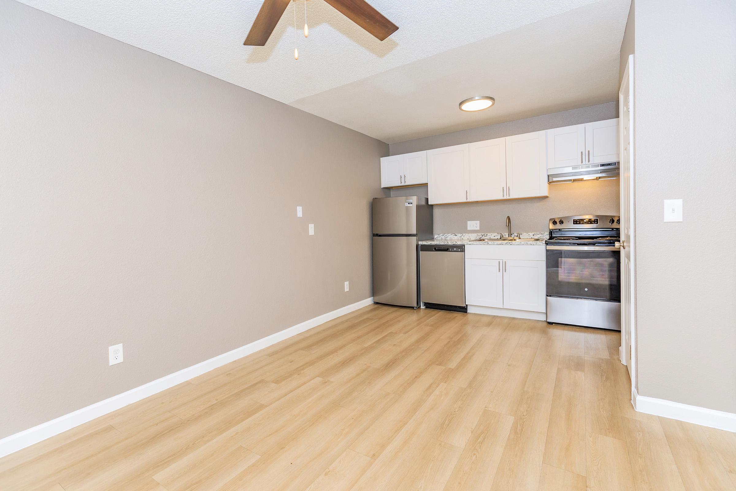 Empty interior of an apartment featuring light-colored walls, a ceiling fan, and laminate flooring. A compact kitchen area with white cabinets, a stainless steel refrigerator, and an oven is visible in the background. The space has good natural light and is ready for furnishing.