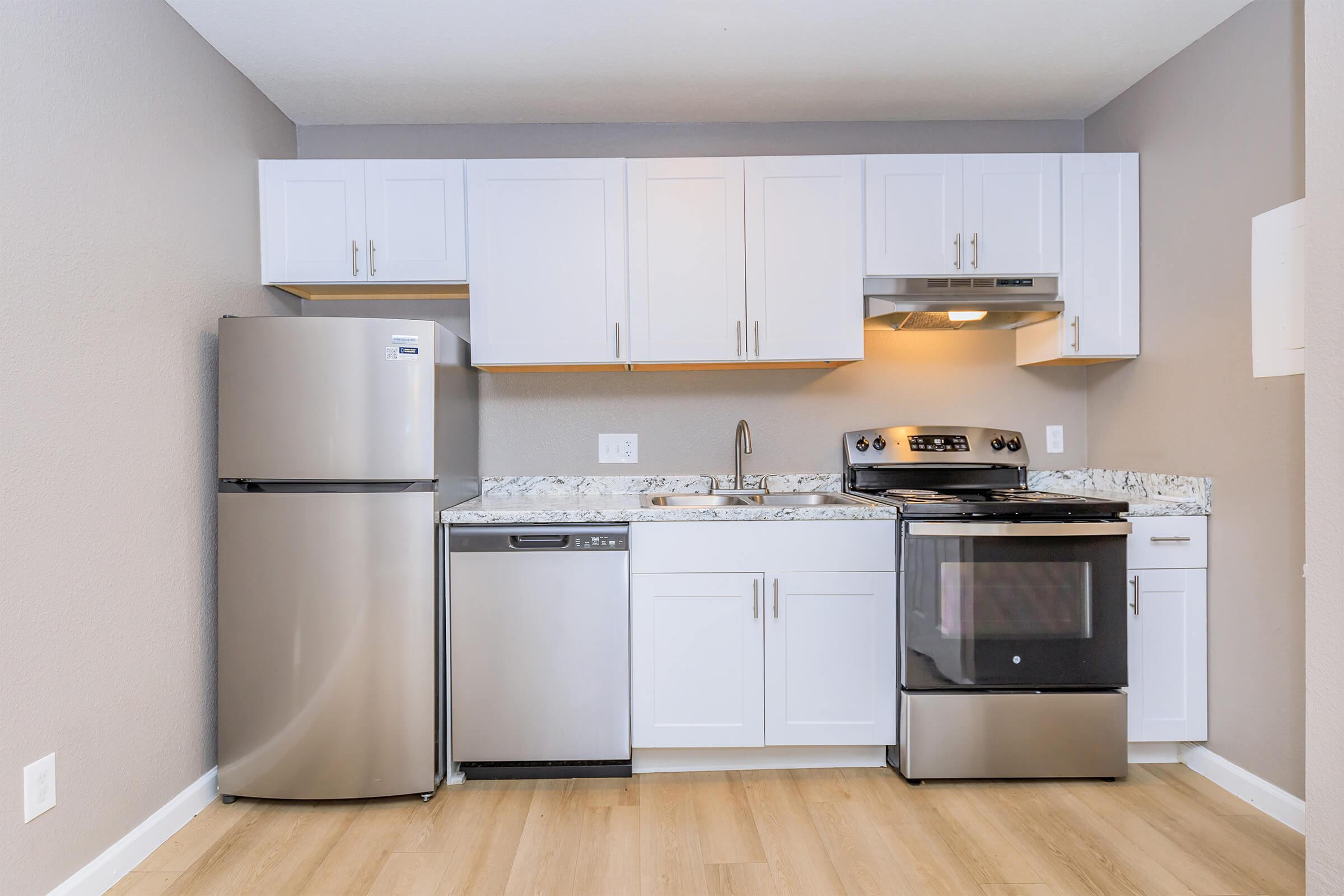 Modern kitchen featuring stainless steel appliances, including a refrigerator, dishwasher, and stove. The cabinets are white with a light gray wall, and the countertops are a patterned granite. The flooring is light-colored, creating a bright and open feel.