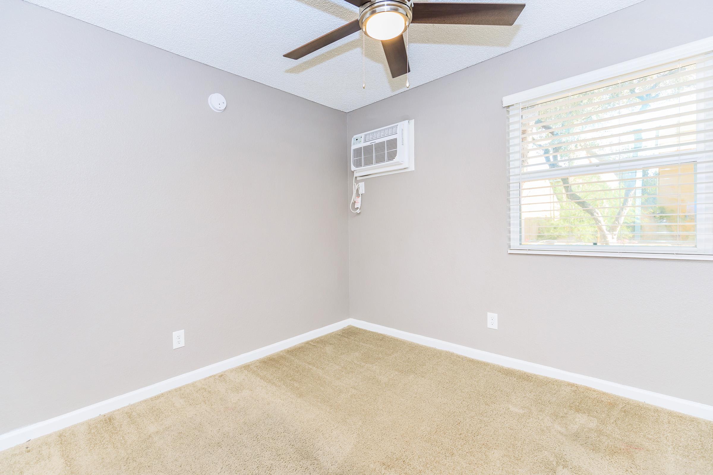 Empty room with light gray walls and a tan carpet. It features a ceiling fan with five blades and an air conditioning unit mounted on the wall. A window with blinds allows natural light to enter, showcasing a view of trees outside. The space is clean and uncluttered, suitable for various purposes.