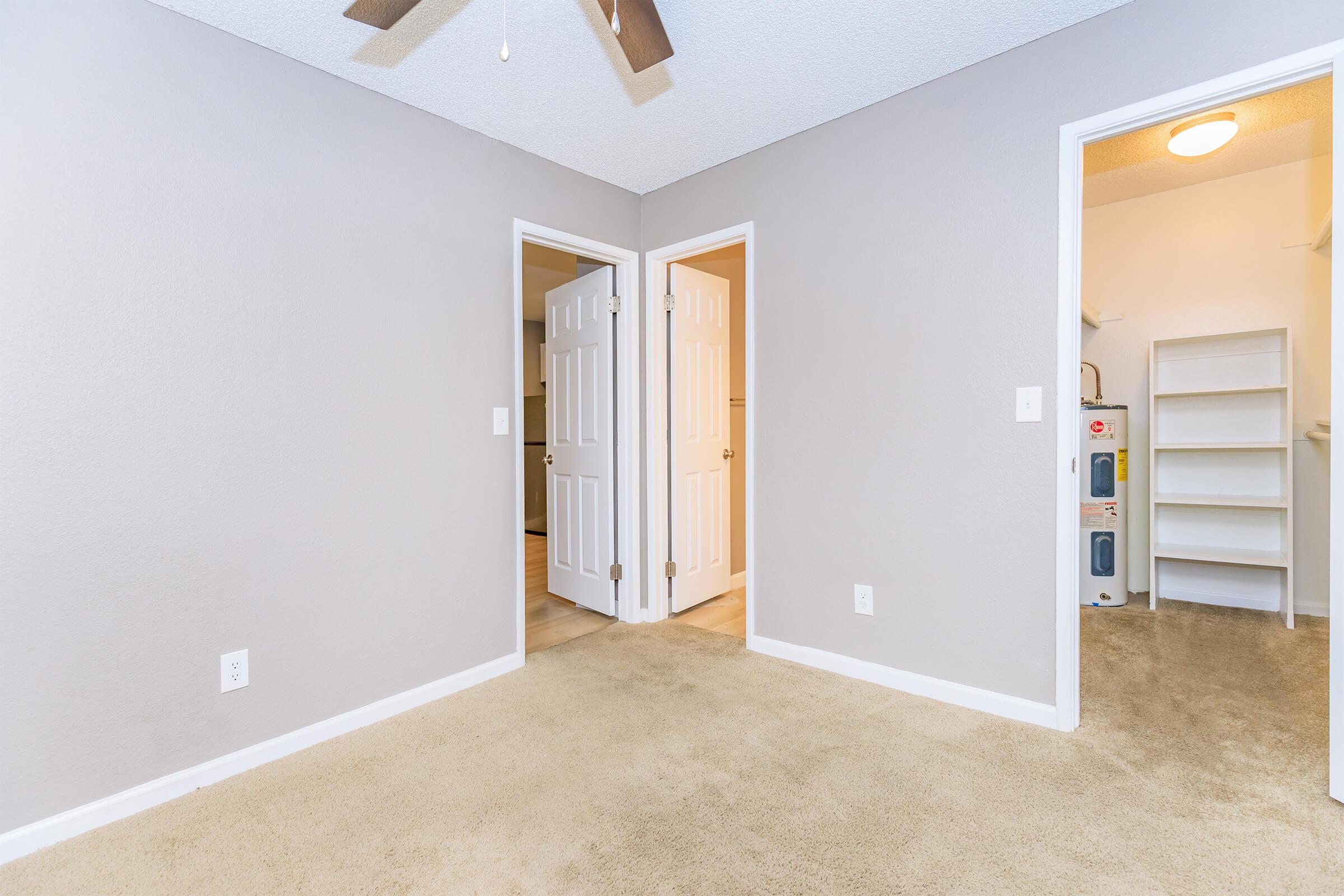 A vacant room with a light beige carpet and gray walls. Two doors lead to adjacent spaces, and there is a light ceiling fan. A doorway on the right opens to a small utility area with shelves, while the door on the left likely leads to another room. Natural light fills the space.