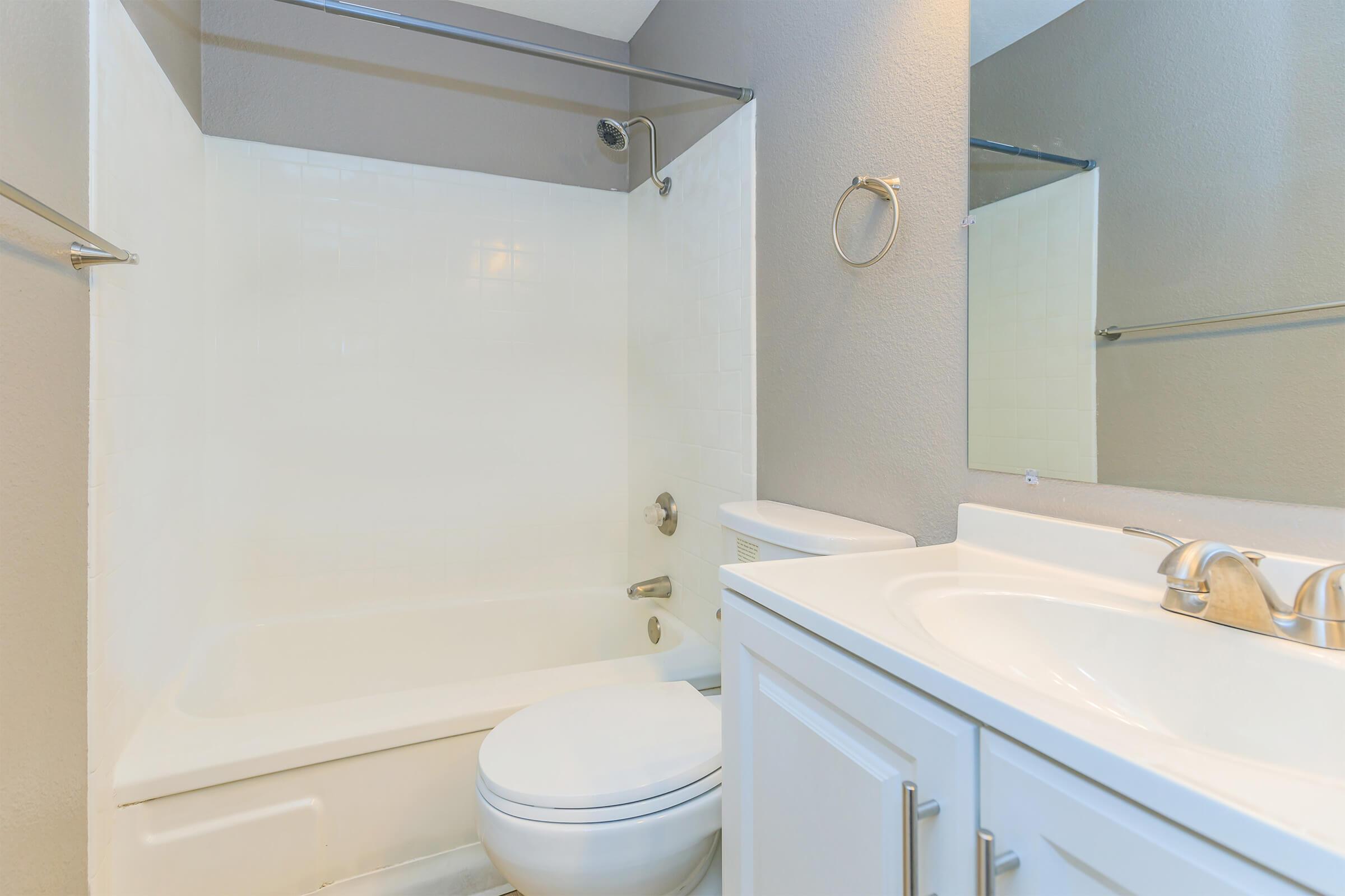 A clean, modern bathroom featuring a shower-tub combo, white cabinetry with a countertop, a single sink, and a large mirror. The walls are painted in a light gray with a neutral tile floor, creating a bright and spacious feel.
