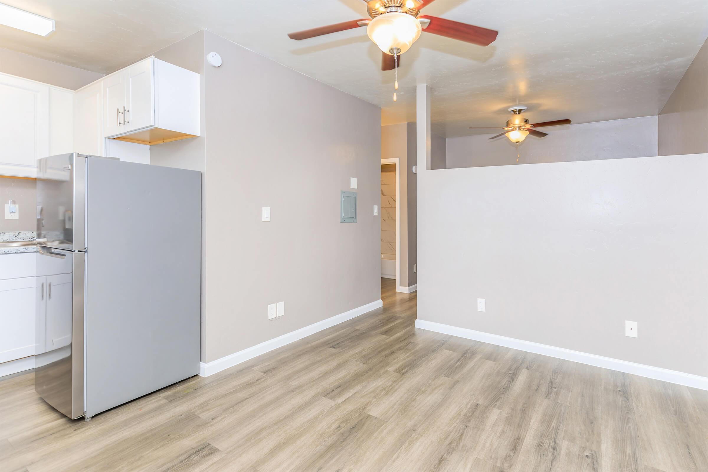 A modern, compact apartment interior featuring a kitchen area with a silver refrigerator, white cabinetry, and a spacious living area. The room has light-colored walls, wooden flooring, and two ceiling fans. A doorway leads to another room at the end of the space.
