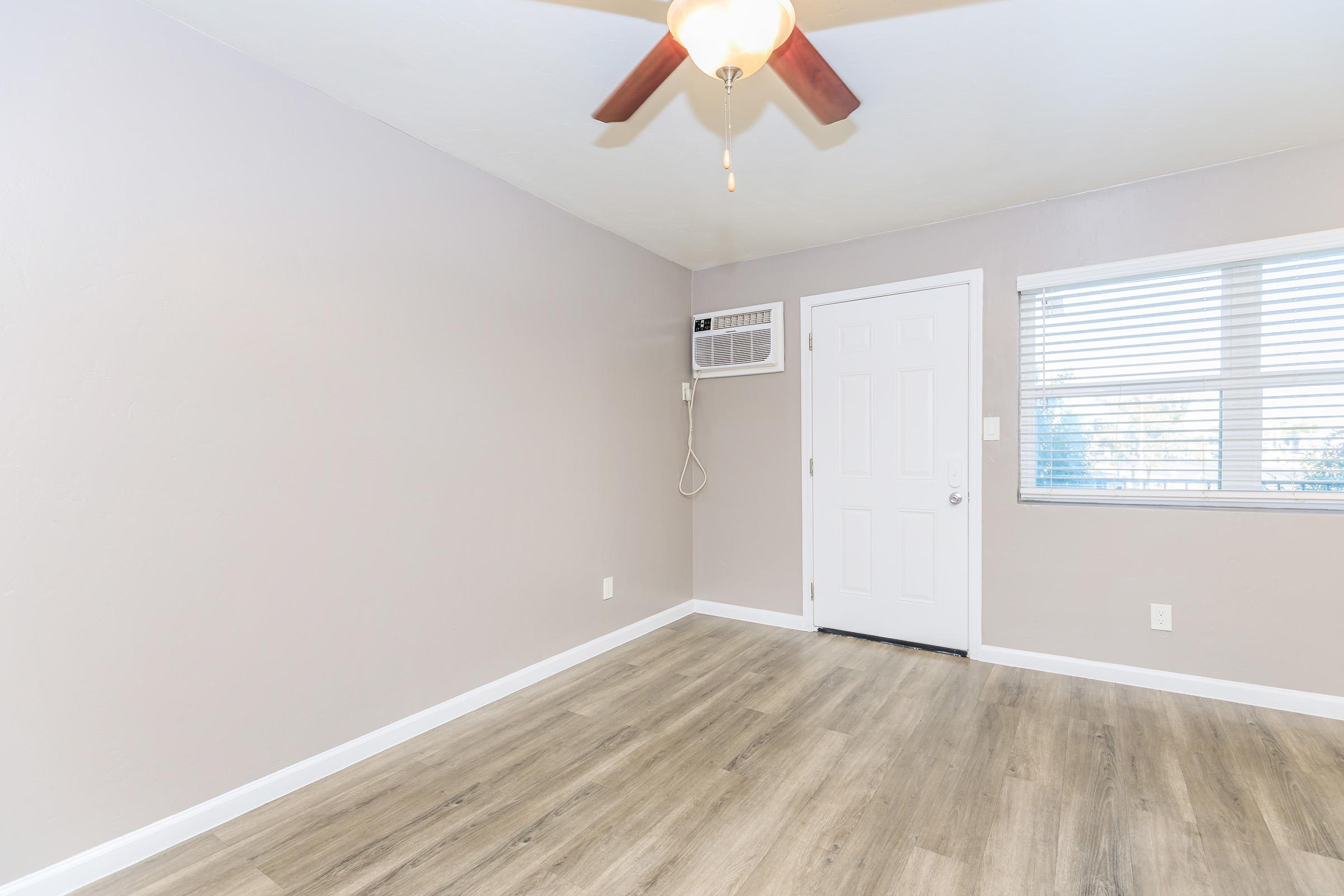 A spacious room with light gray walls, a ceiling fan, and a hardwood floor. It features a door with a window next to it, allowing natural light to enter. An air conditioning unit is mounted on the wall. The overall ambiance is bright and clean, suitable for various uses.