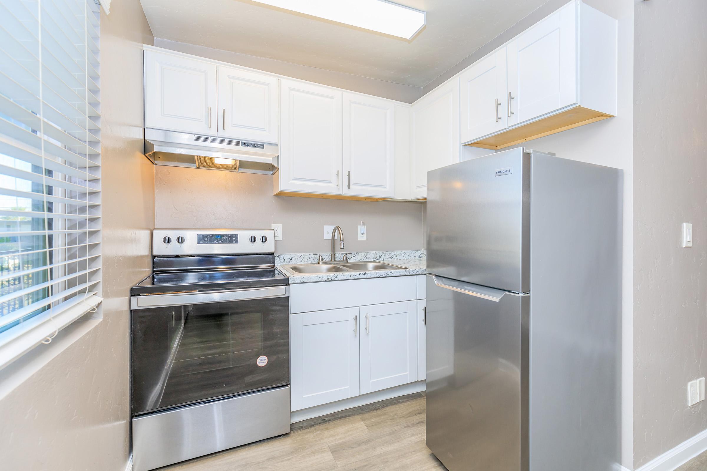 Modern kitchen featuring white cabinetry, stainless steel appliances including a refrigerator and oven, and a double sink with a marble countertop. Natural light streams in through a window with white blinds, enhancing the spacious and clean design.