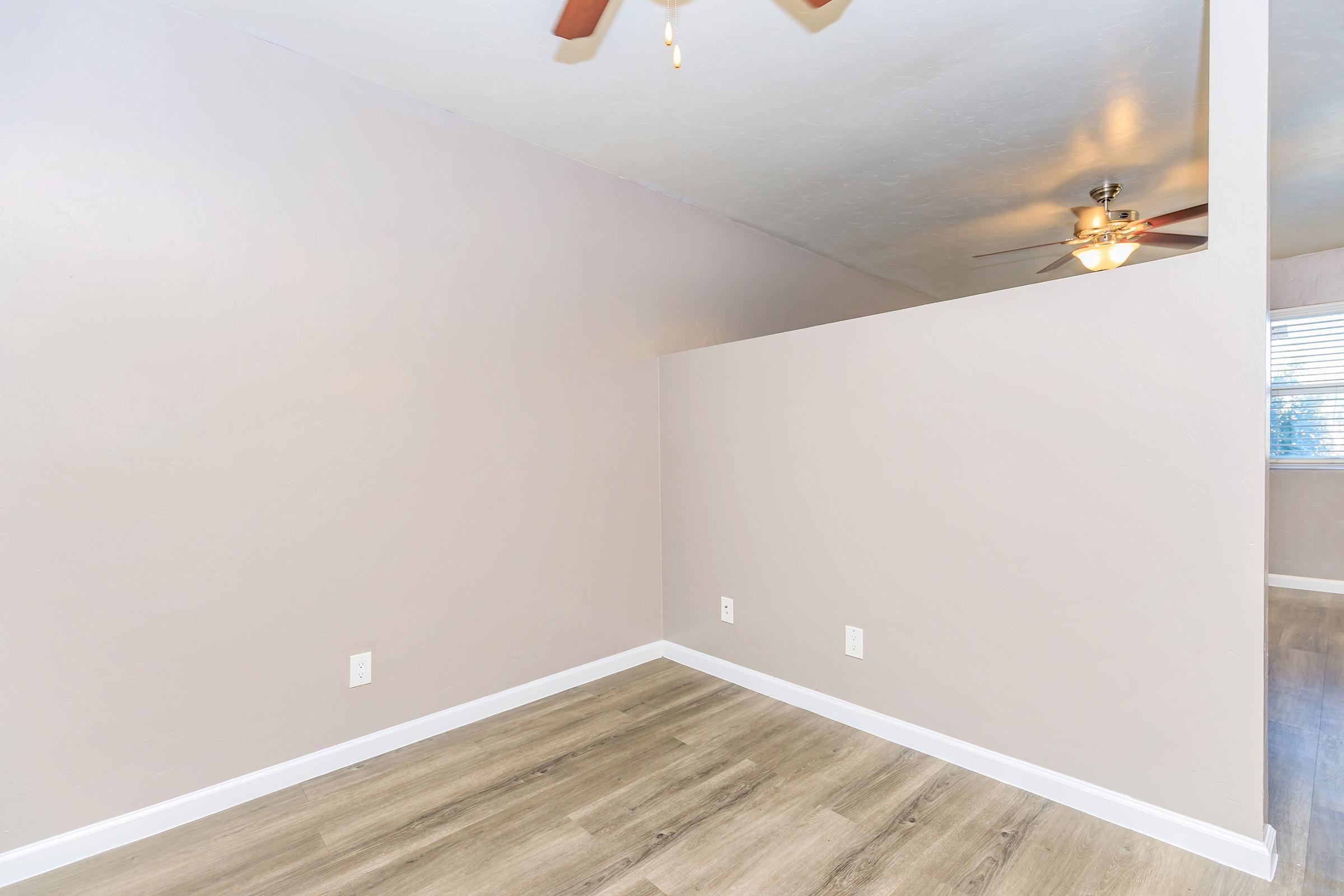A minimalistic interior view of a room featuring light-colored walls, a ceiling fan, and wooden flooring. There is a partition wall creating an open yet divided space, and a window is visible in the background, allowing natural light to enter.