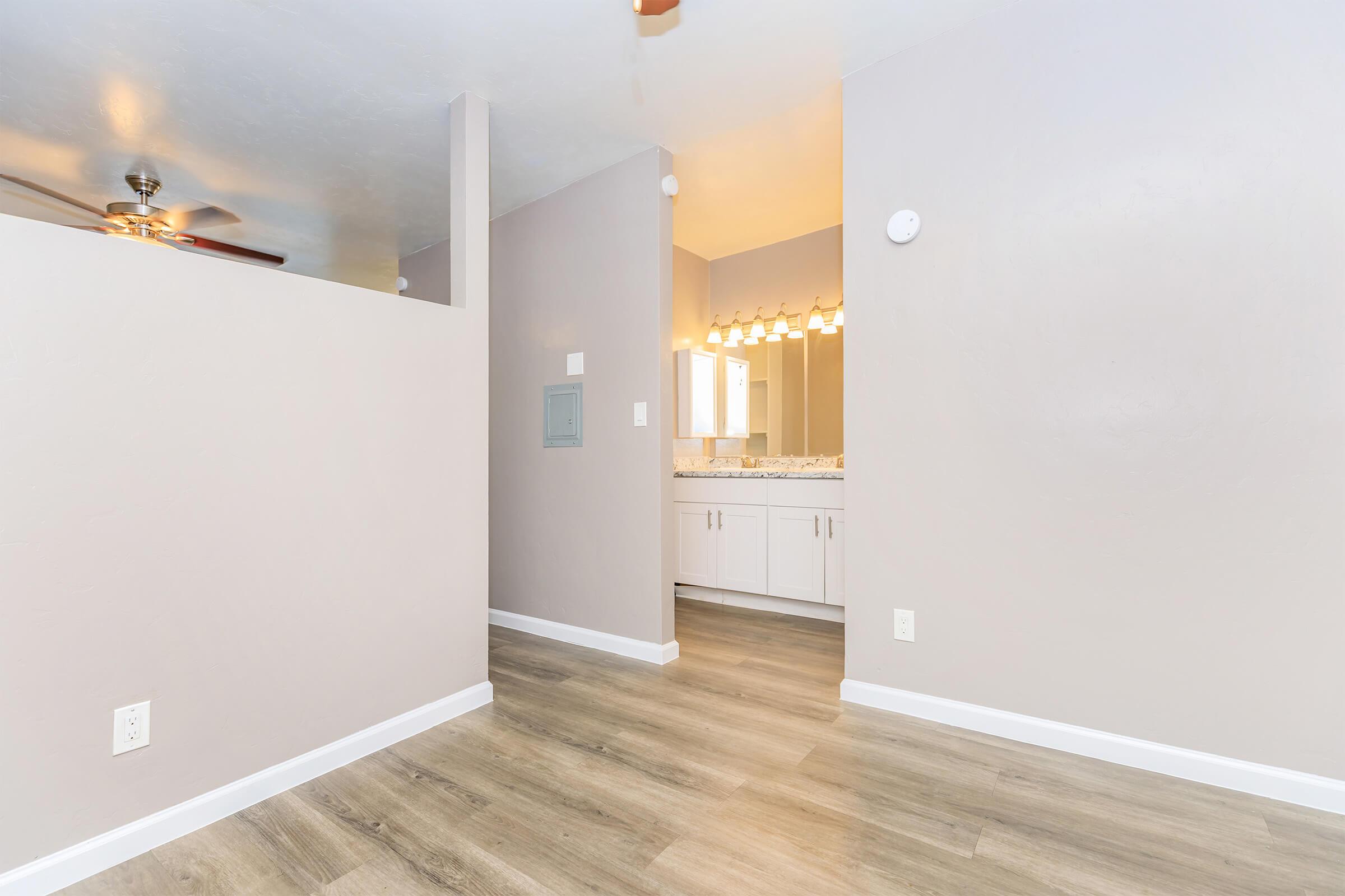 A view of a modern, light-colored interior space featuring wooden flooring, a partial wall, and a bathroom area with a vanity and lighting fixtures. The room has a neutral color palette, and a ceiling fan is visible in the background.