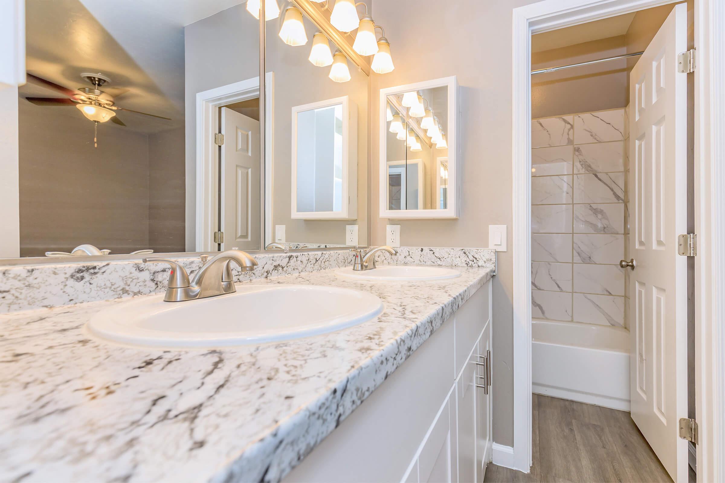 A modern bathroom featuring a dual sink vanity with a marble countertop, a large mirror, and stylish light fixtures. The space includes a shower with marble-like tiles and a bathtub, all set against a neutral wall color and sleek flooring.