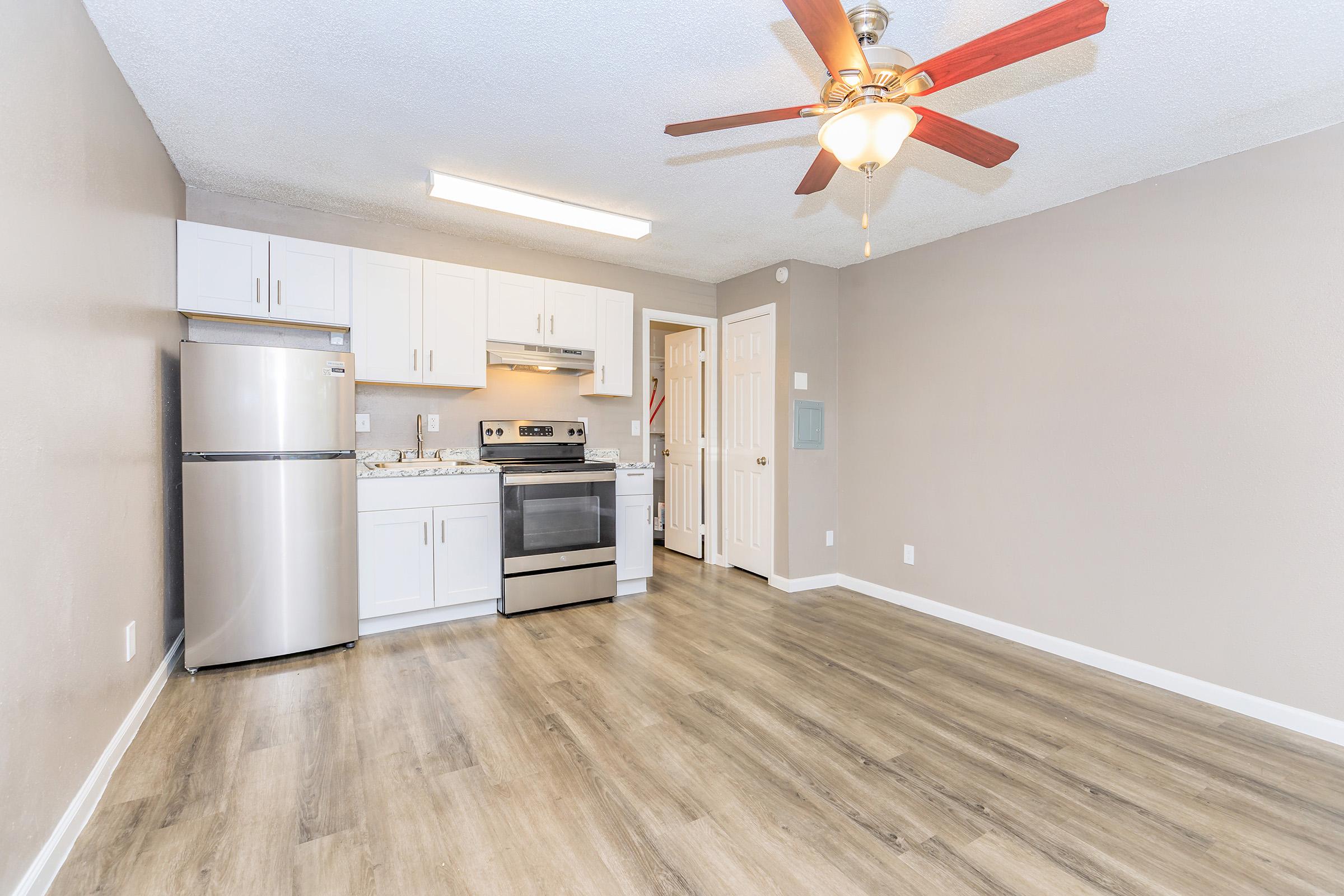 A modern, bright kitchen and living area featuring light-colored walls, a ceiling fan with red blades, stainless steel appliances including a refrigerator and stove, and laminate flooring. The space appears clean and is ready for furniture arrangement.