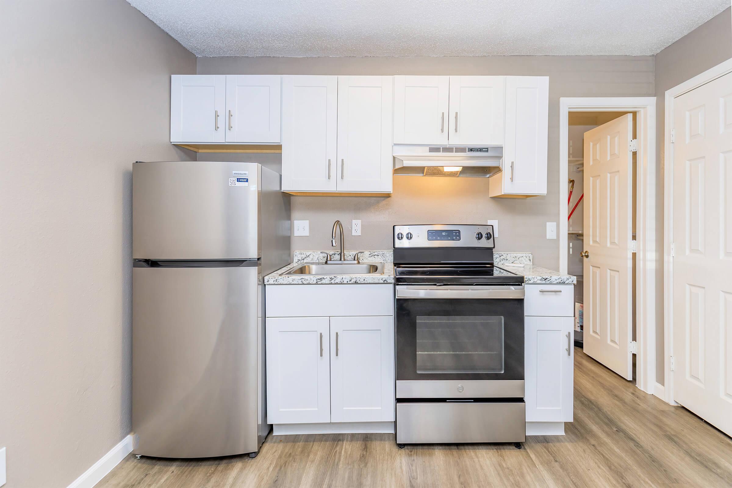 A modern kitchen featuring white cabinets, a stainless steel refrigerator, oven, and a microwave. The countertop is made of speckled stone, and there's laminate flooring. The kitchen is well-lit with natural light coming from the door on the right.