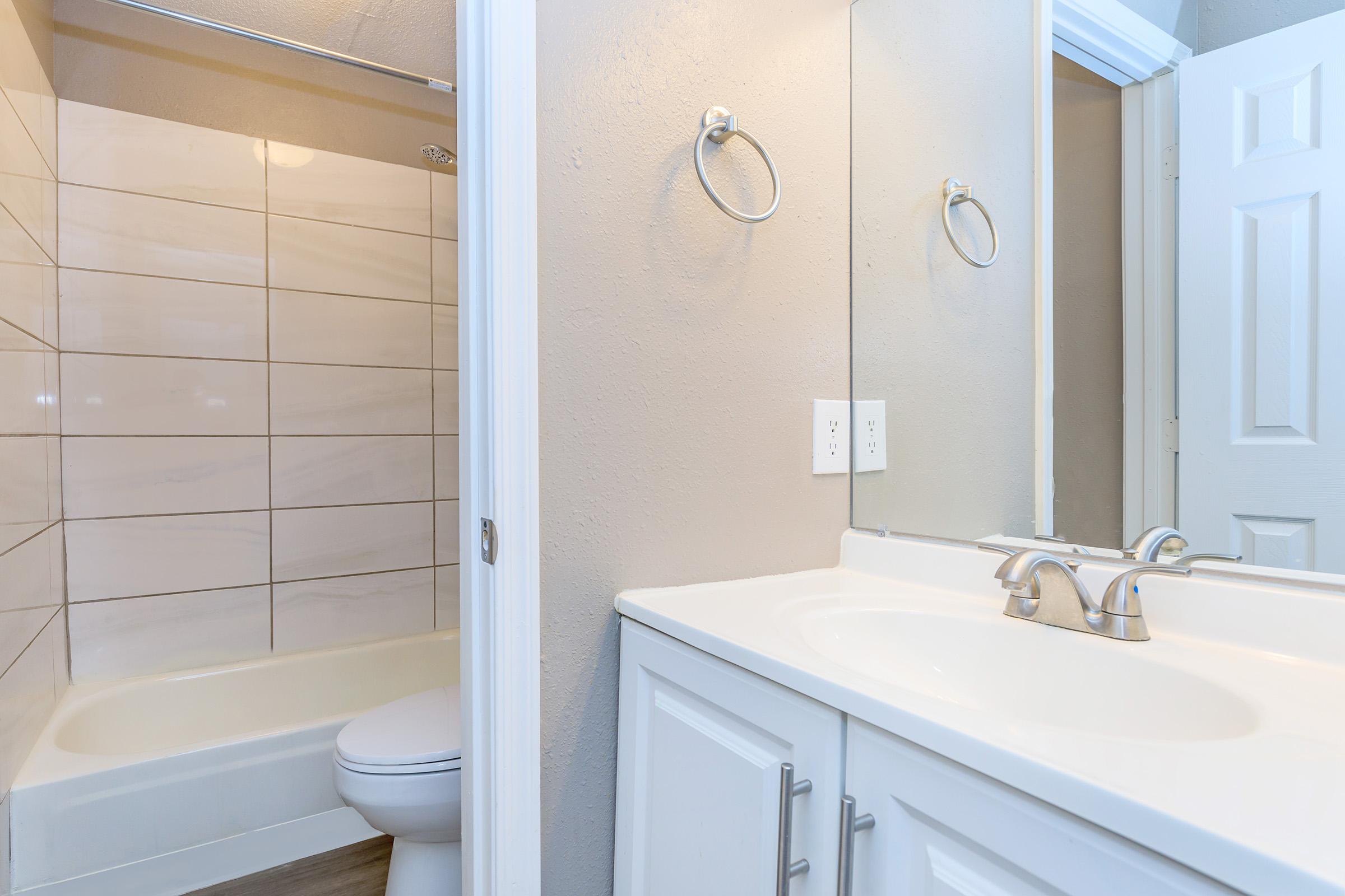 A clean and modern bathroom featuring a white vanity with dual sinks, a large mirror, and a towel ring on the wall. In the background, a shower/tub combo is visible with tiled walls. The space is bright and well-lit, showcasing a contemporary design.