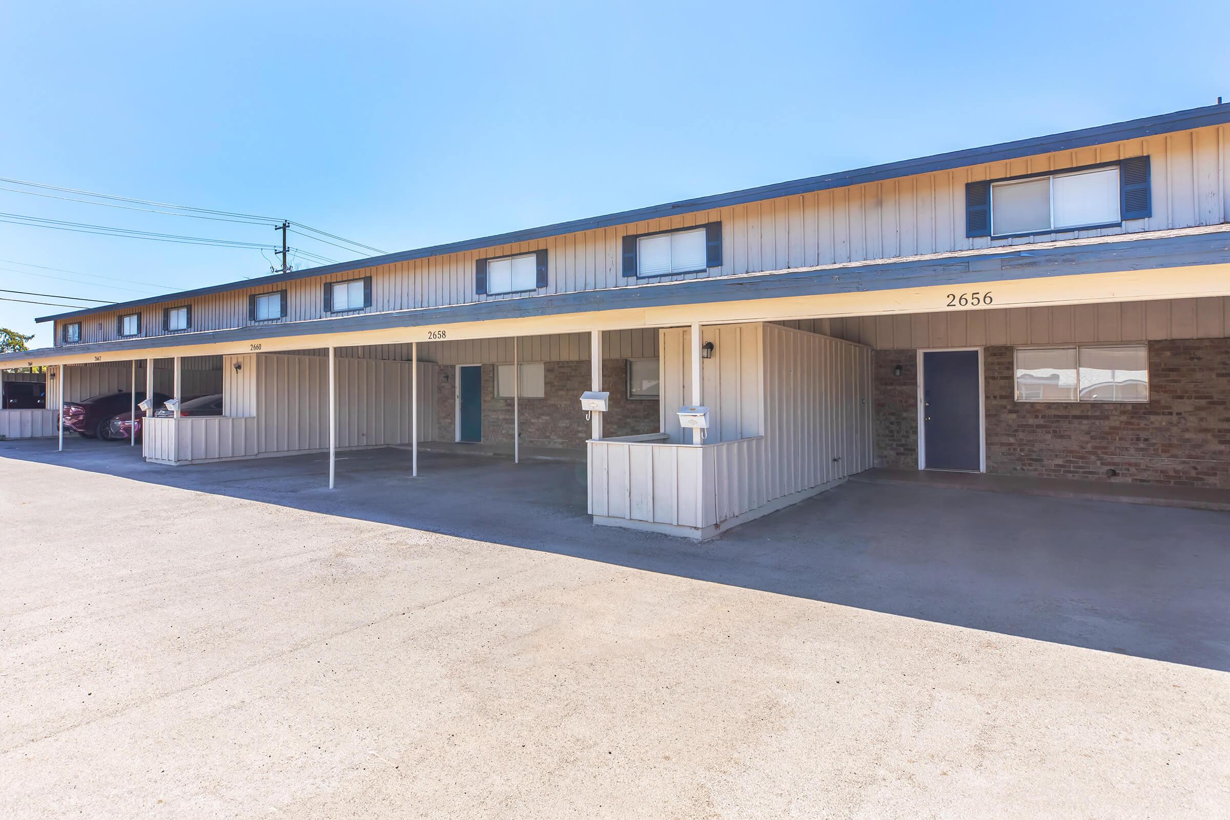 A row of modest apartment units featuring covered parking spaces beneath a two-story building with light-colored siding. Each unit has a front door, and mailboxes are mounted on the wall. The parking lot is paved and features clear blue skies in the background.