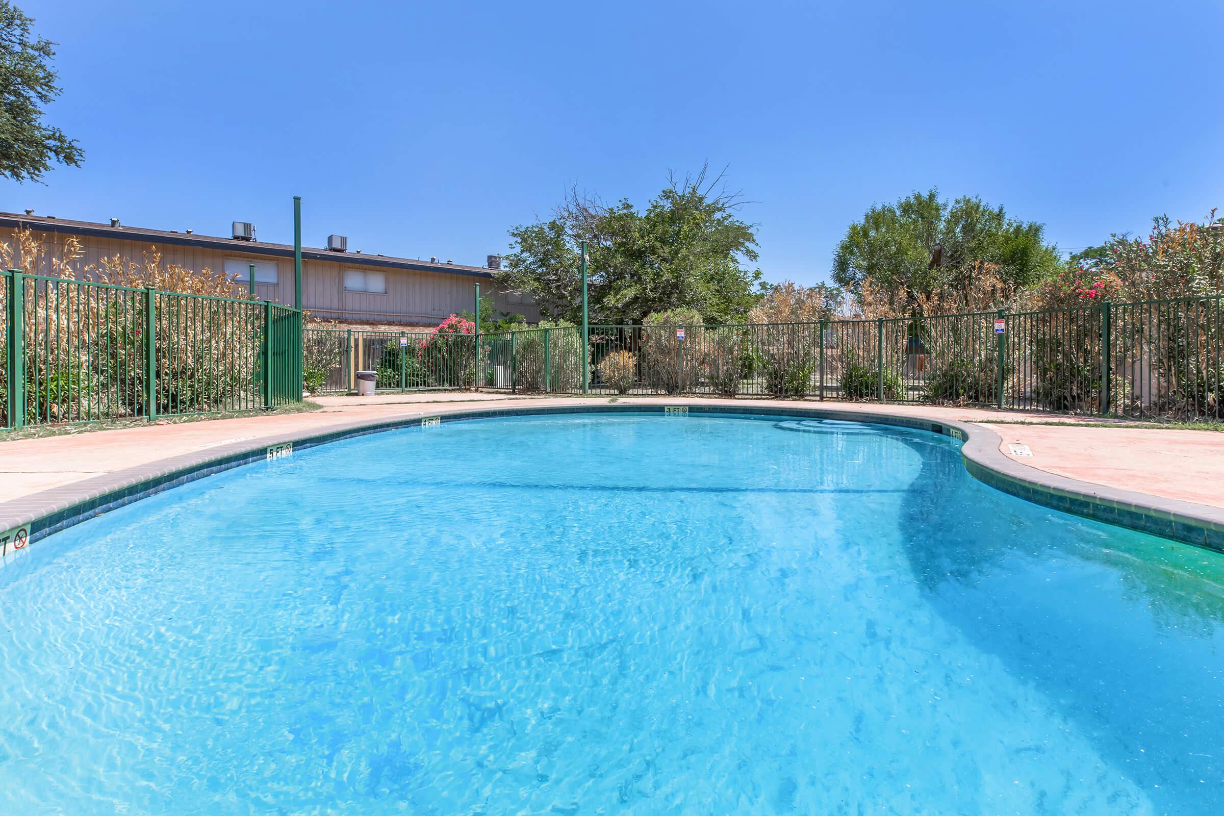 A clear swimming pool surrounded by a green fence, with lush plants and trees in the background. The sky is bright blue, indicating a sunny day. The pool's water appears inviting and calm.