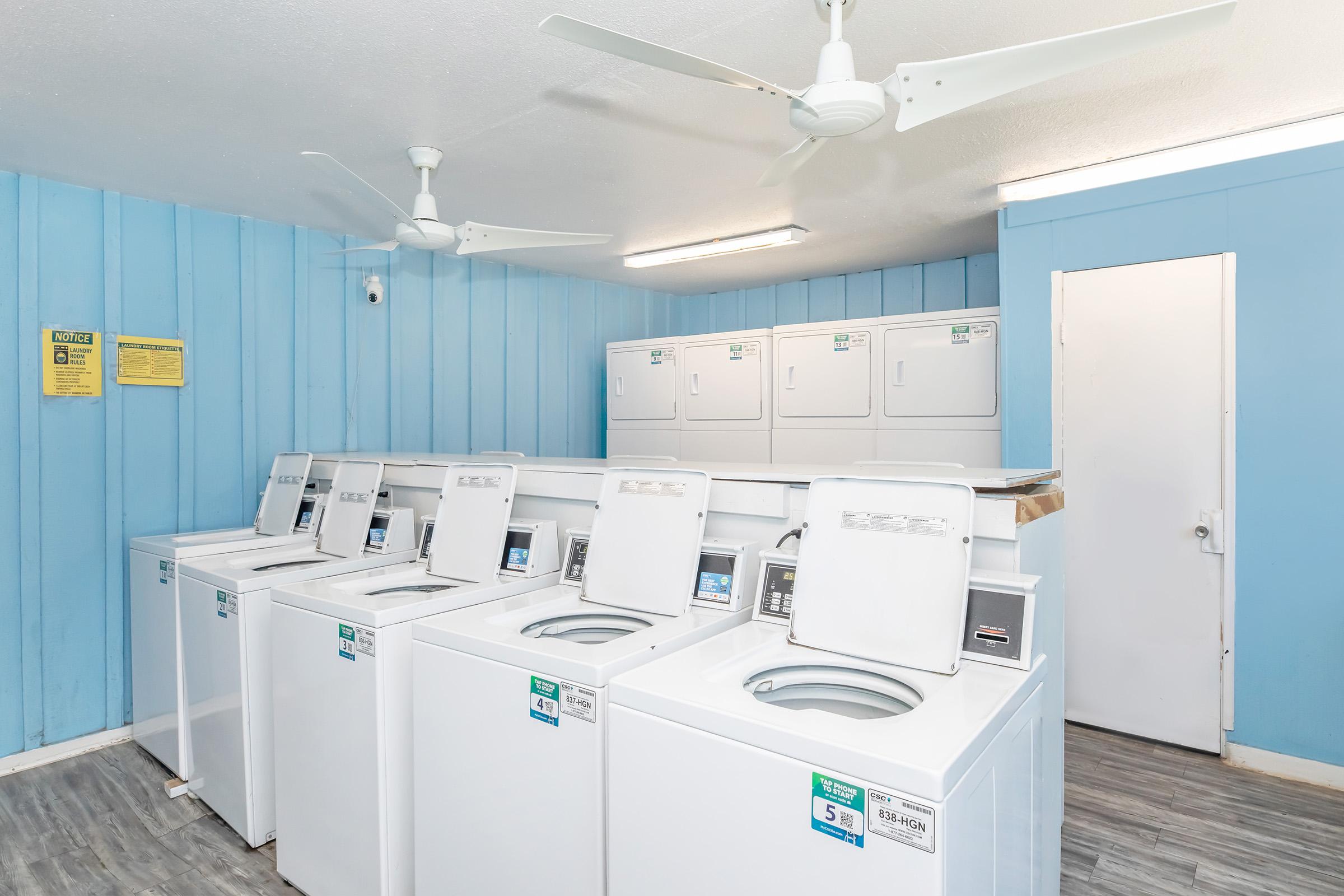 A bright laundry room featuring several white washing machines arranged in a row against blue walls. Ceiling fans are installed above for ventilation. There are also laundry dryers in the background and a door leading to another area. Informational signs are visible on the walls.