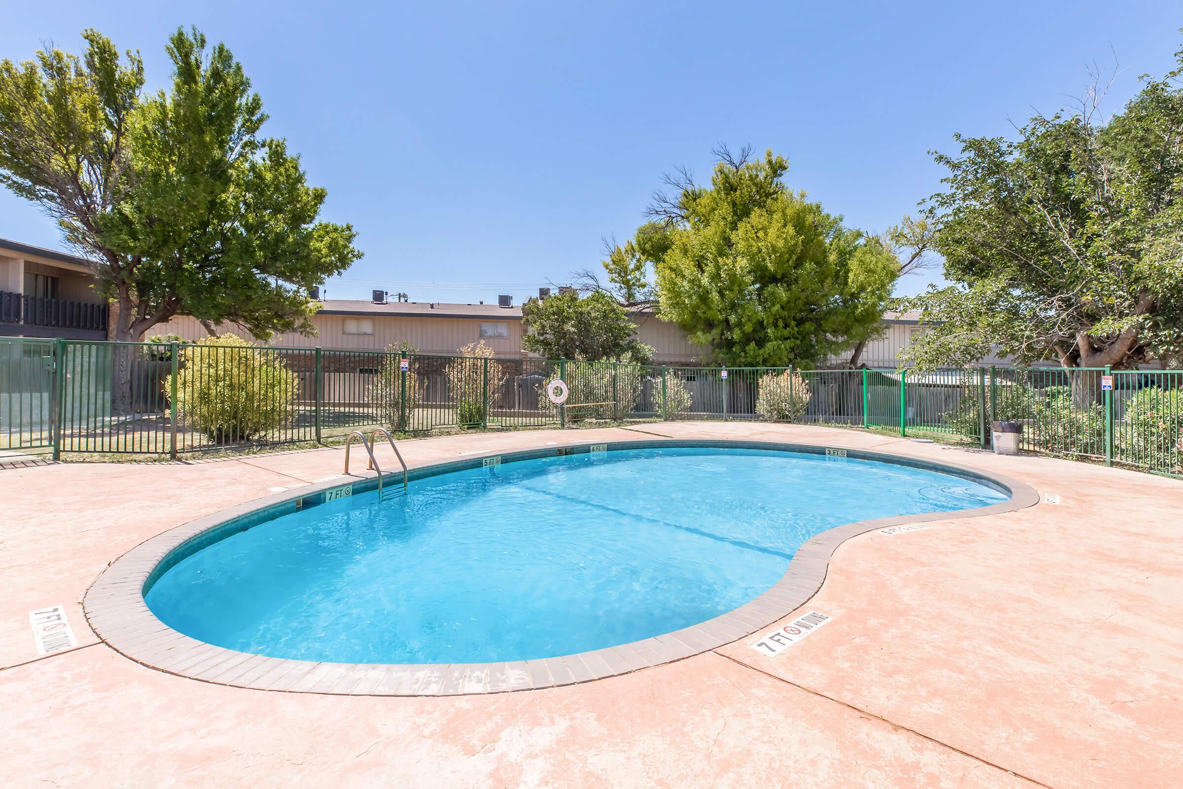 A clear swimming pool surrounded by a concrete deck, with green trees and shrubs in the background. A fence encloses the pool area, and the sky is bright blue. The pool features a ladder for entry and a depth marker visible on the edge. The overall scene conveys a relaxing outdoor atmosphere.
