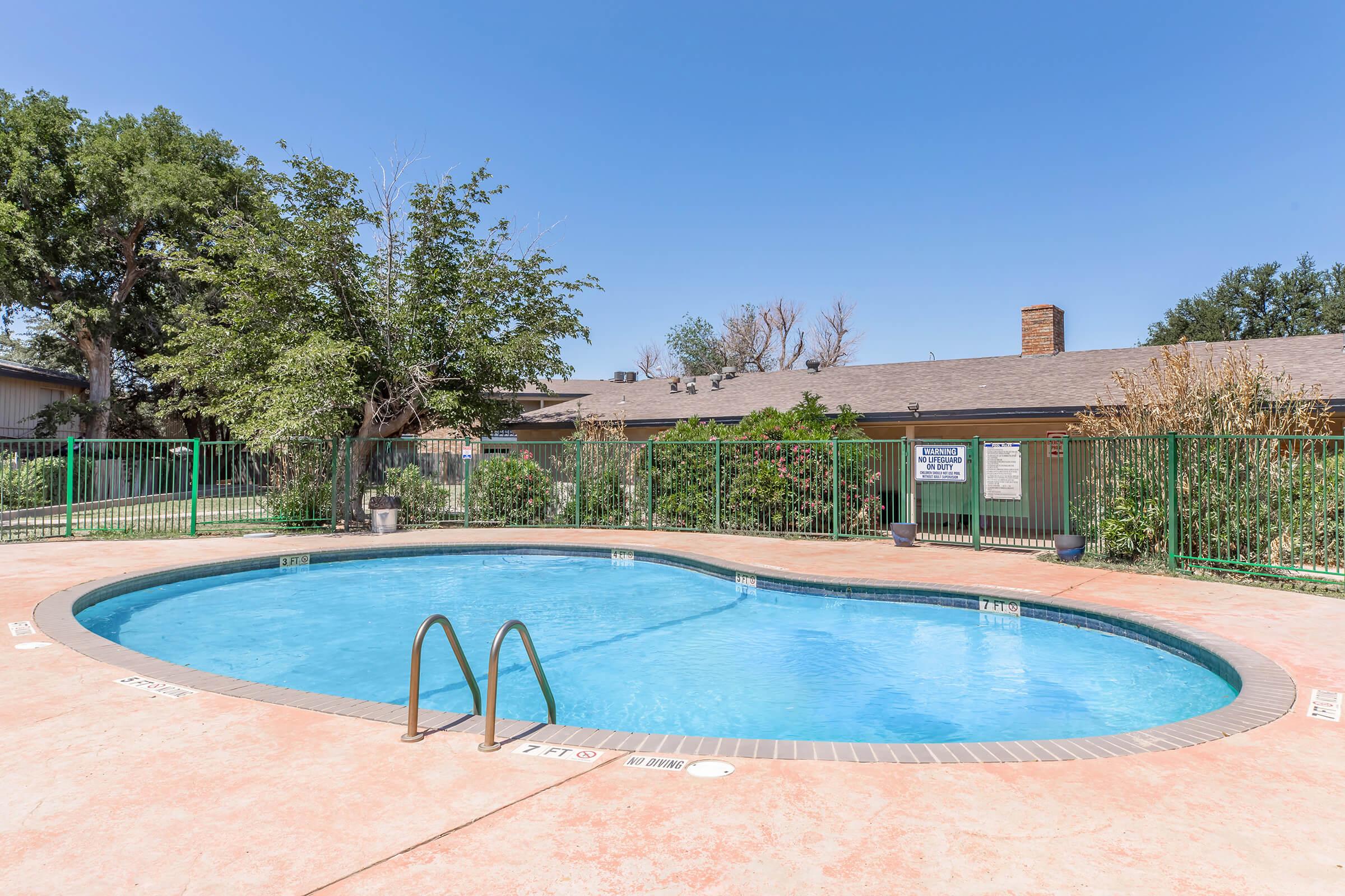 A clear blue swimming pool surrounded by a green fence, with a small grassy area and trees in the background. The pool has steps for entry, and a building is visible in the distance with vegetation nearby. Blue sky with few clouds overhead.