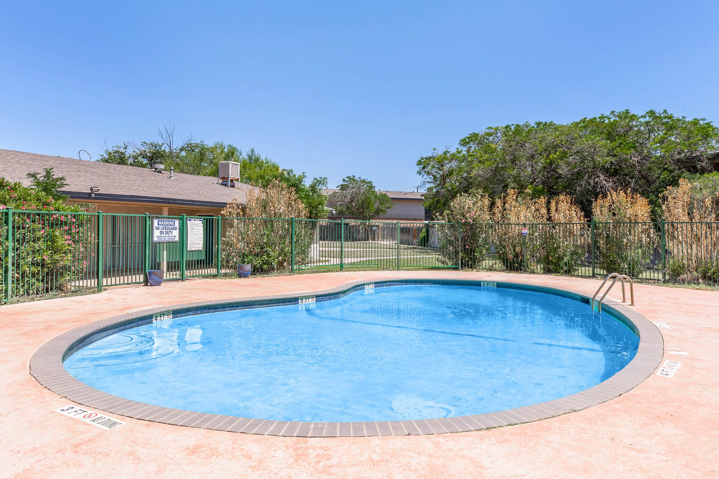 A clear blue swimming pool surrounded by a green fence and lush vegetation on a sunny day. In the background, there is a building with a sign, and the area is well-maintained with decorative plants and grass.
