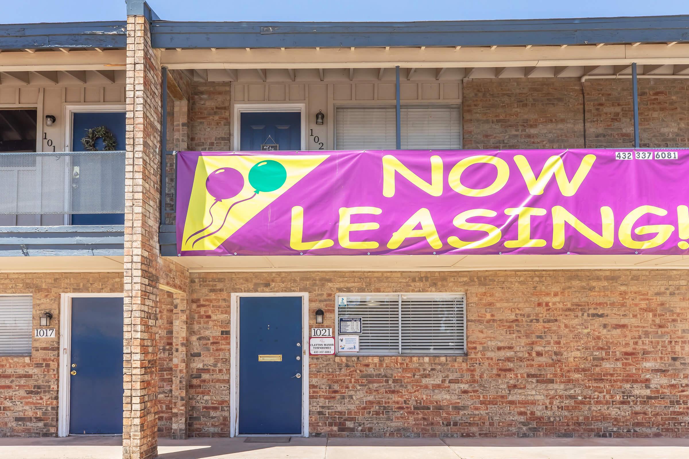 Bright purple banner on a brick building displaying "NOW LEASING!" with colorful balloons. Below the banner, there are multiple apartment doors, some with numbers visible. The setting appears to be a multi-unit residential complex.