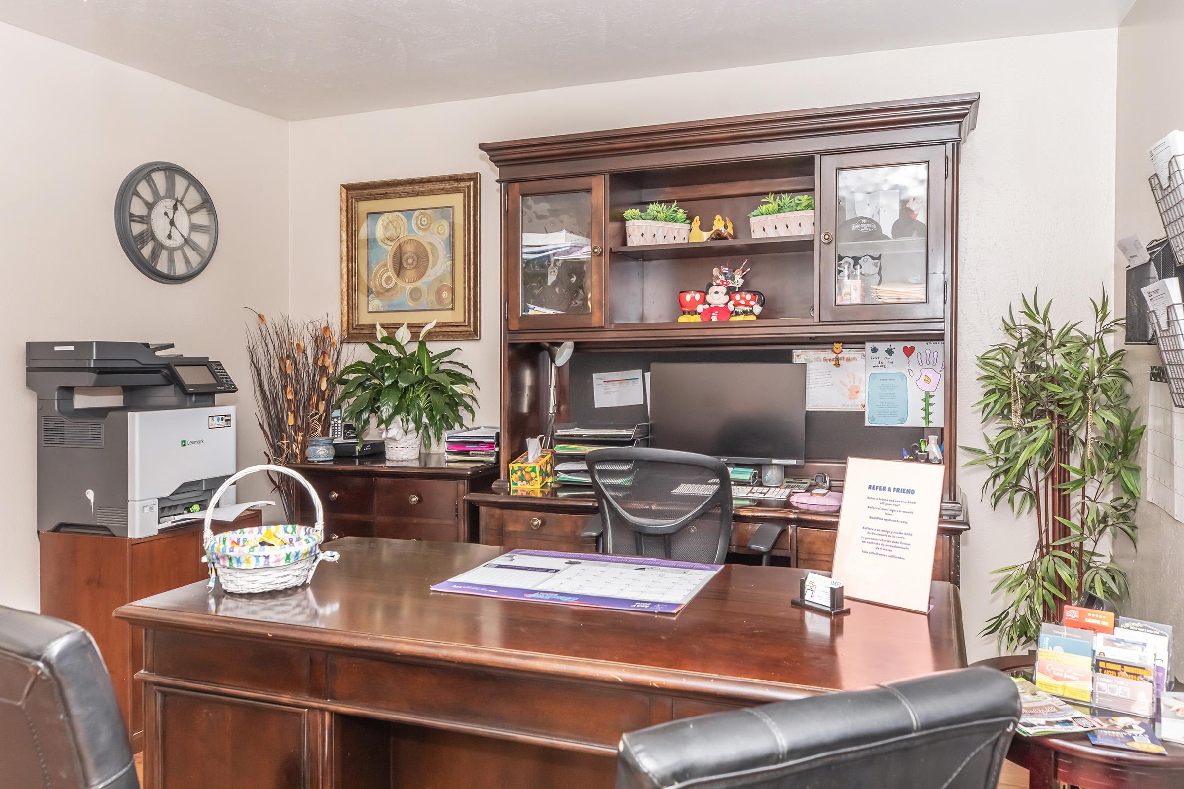 A modern home office featuring a large wooden desk with a calendar and a decorative basket. A printer is on the left side, and the wall displays a clock and framed artwork. Plants decorate the space, alongside shelves with small potted plants and other items. The overall atmosphere is organized and inviting.
