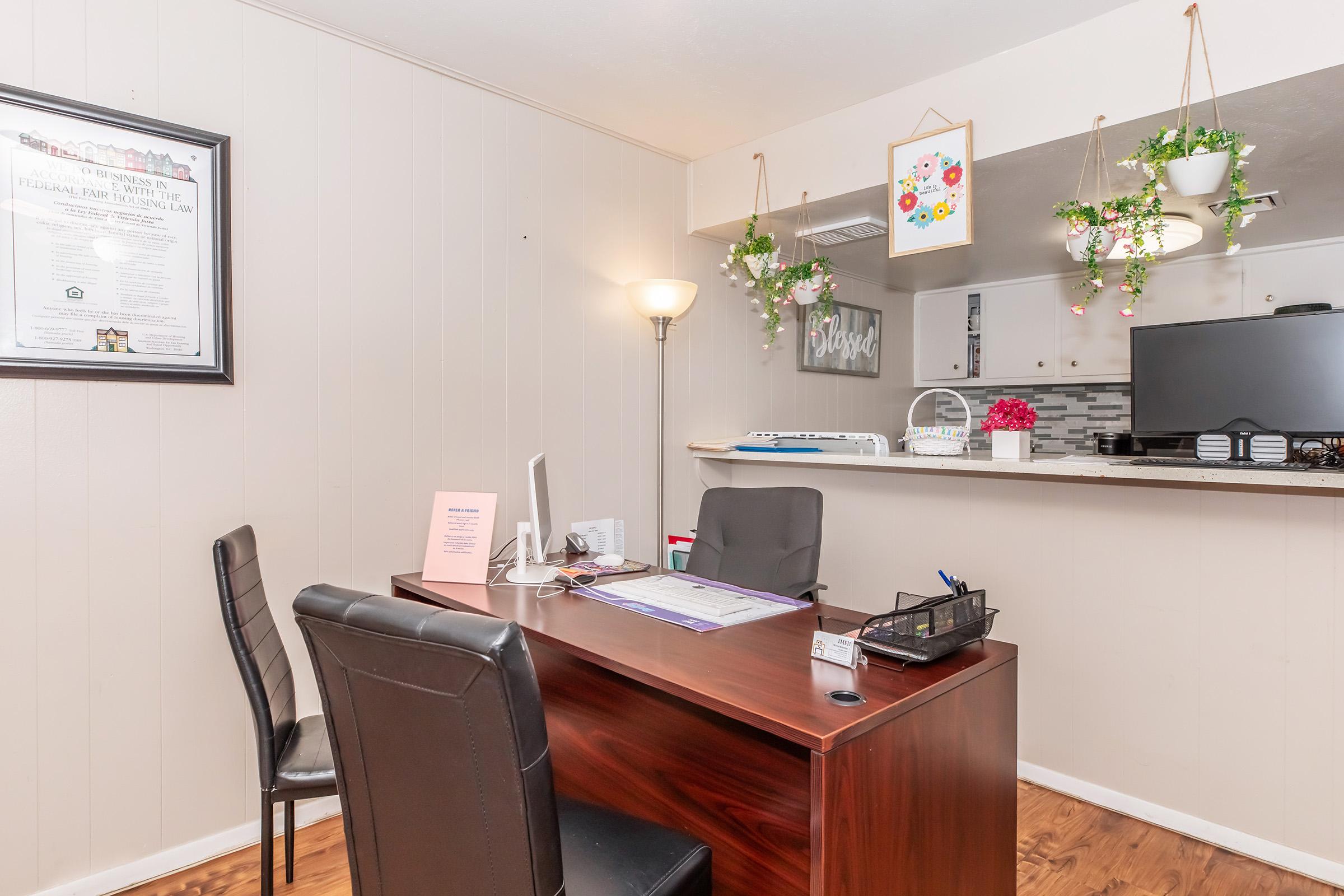 A small office space featuring a wooden desk with two black chairs, a computer, and paperwork. On the wall hangs a framed document. The area has indoor plants and decorations, with a view of a kitchen area in the background. Bright and inviting atmosphere.