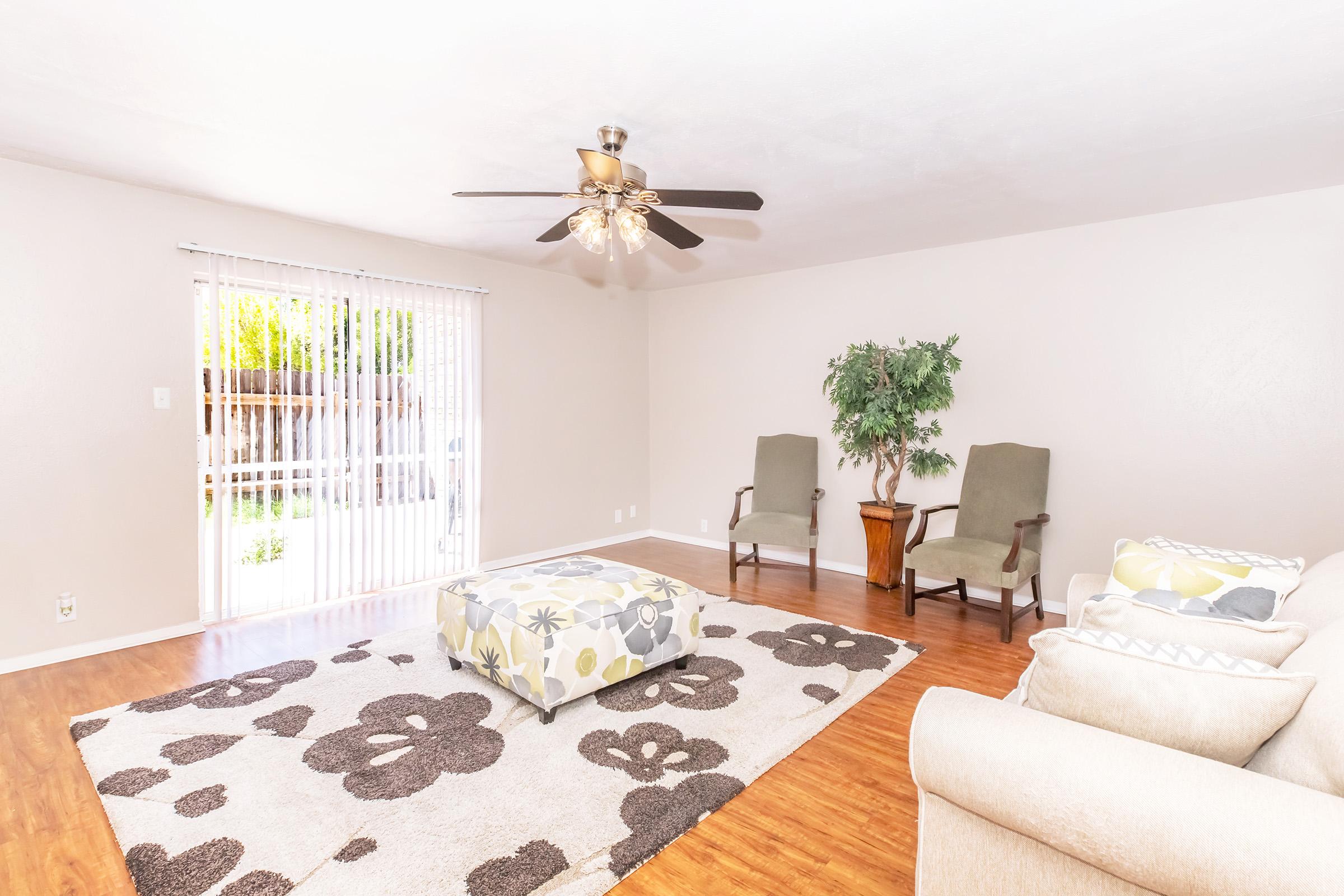 A spacious living room featuring light-colored walls, a ceiling fan with lights, two green armchairs, a potted plant, and a large area rug with floral patterns. A coffee table with a patterned top sits on the rug, and large sliding glass doors allow natural light and views of the outside greenery.
