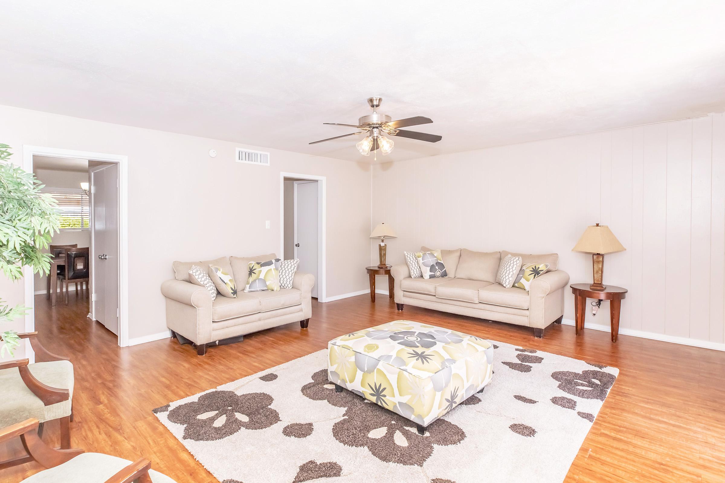 A light, airy living room featuring two beige sofas with decorative pillows, a patterned ottoman in the center, and a large area rug with floral designs. A ceiling fan hangs above, and there are two doorways leading to other rooms. Natural light fills the space, creating a welcoming atmosphere.