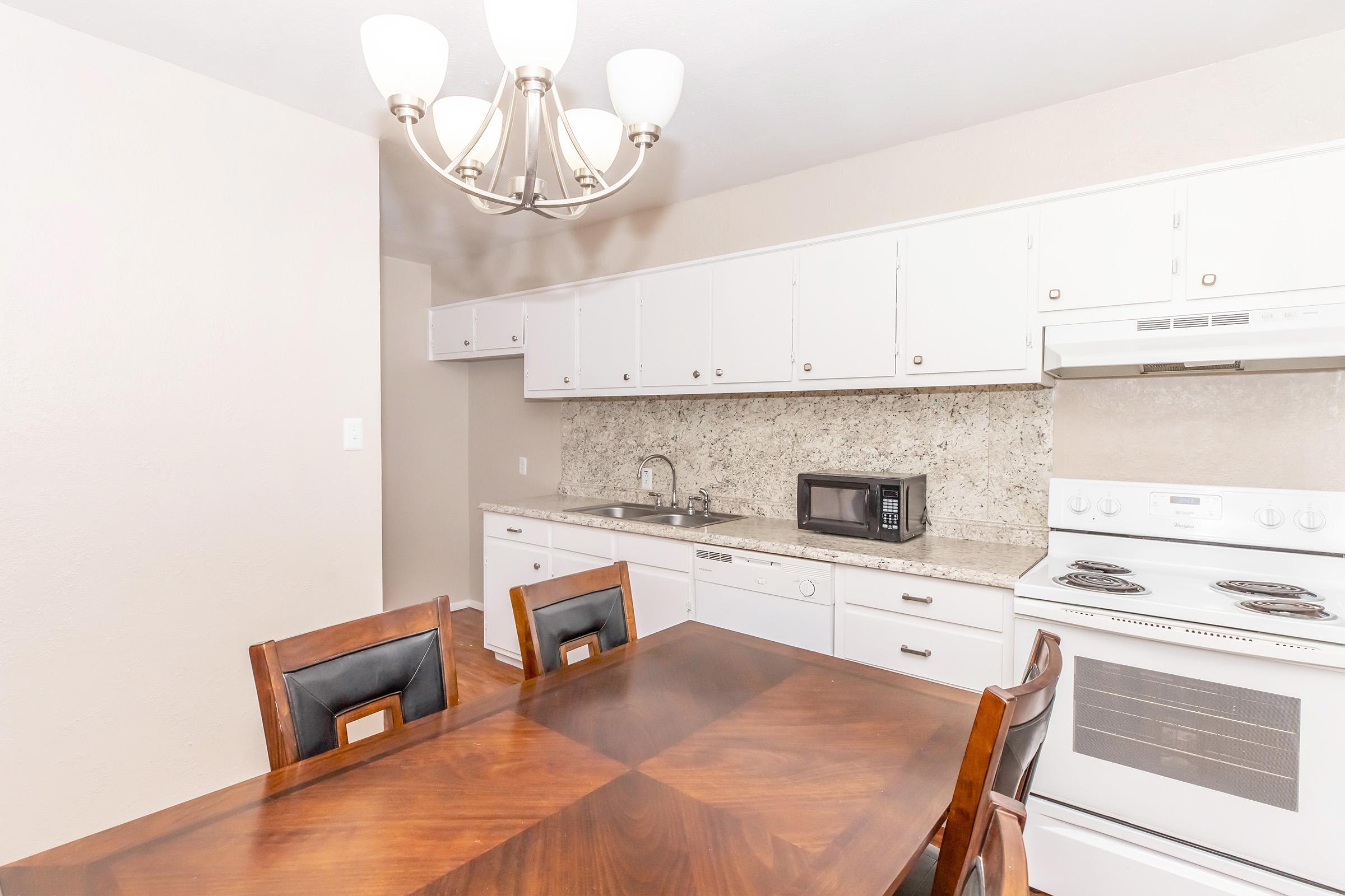 A bright, modern kitchen featuring white cabinetry, a granite countertop, and stainless steel appliances. The image shows a dining table with dark wood chairs, a microwave on the countertop, and a ceiling light fixture above. The overall atmosphere is clean and inviting.