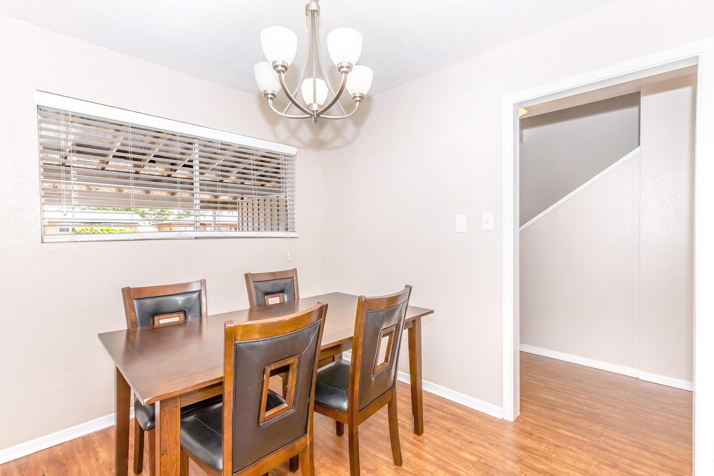 A well-lit dining area featuring a wooden table with four black chairs, a stylish chandelier overhead, and a window with blinds. A doorway leads to a staircase, and the walls are painted in a light neutral color, complemented by laminate flooring.