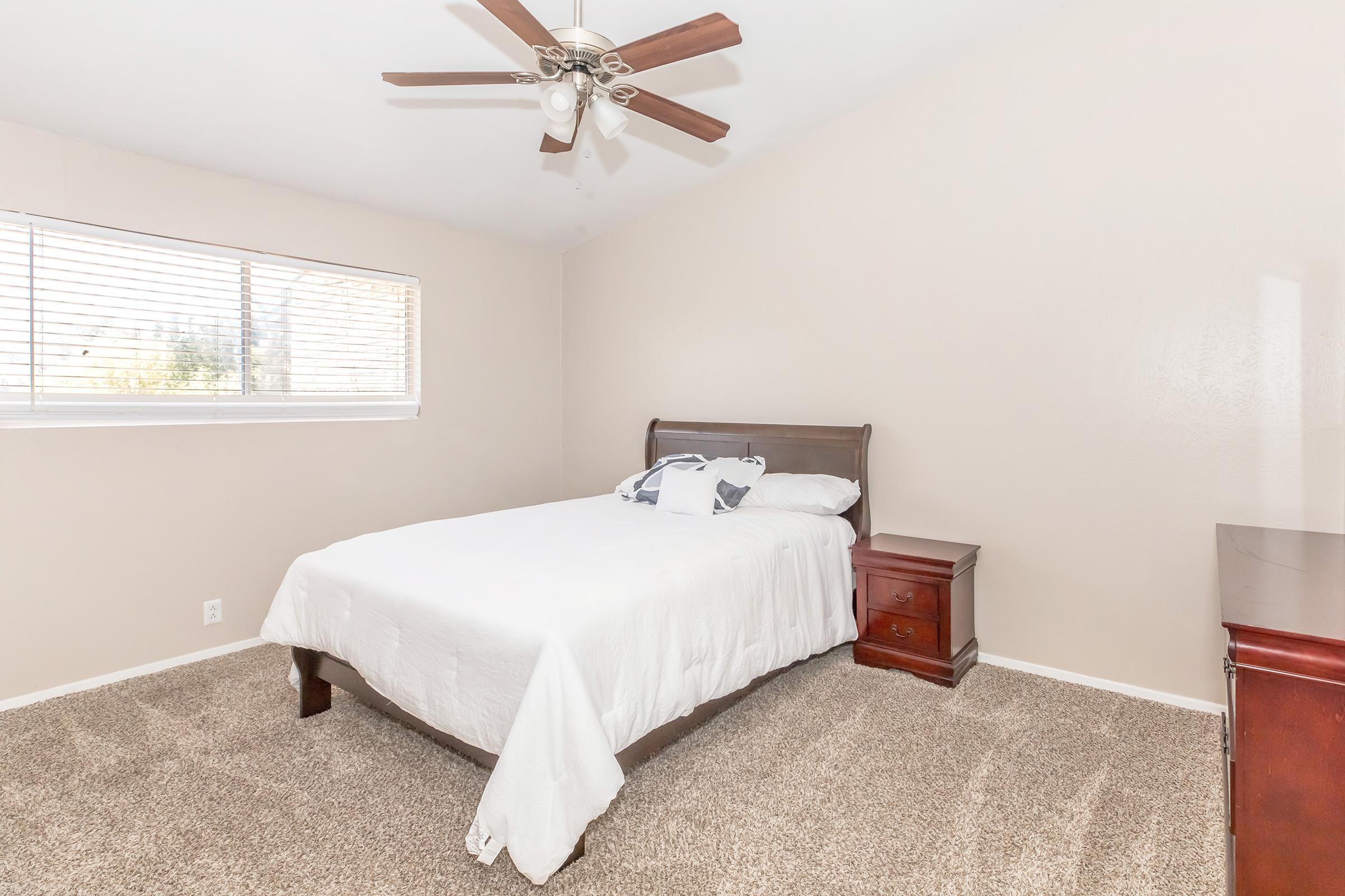 Spacious bedroom featuring a queen-sized bed with a white bedspread, a wooden nightstand, and a light-colored wall. A ceiling fan is visible, and there's a window with blinds allowing natural light. The floor is carpeted in a neutral tone, enhancing the cozy atmosphere of the room.