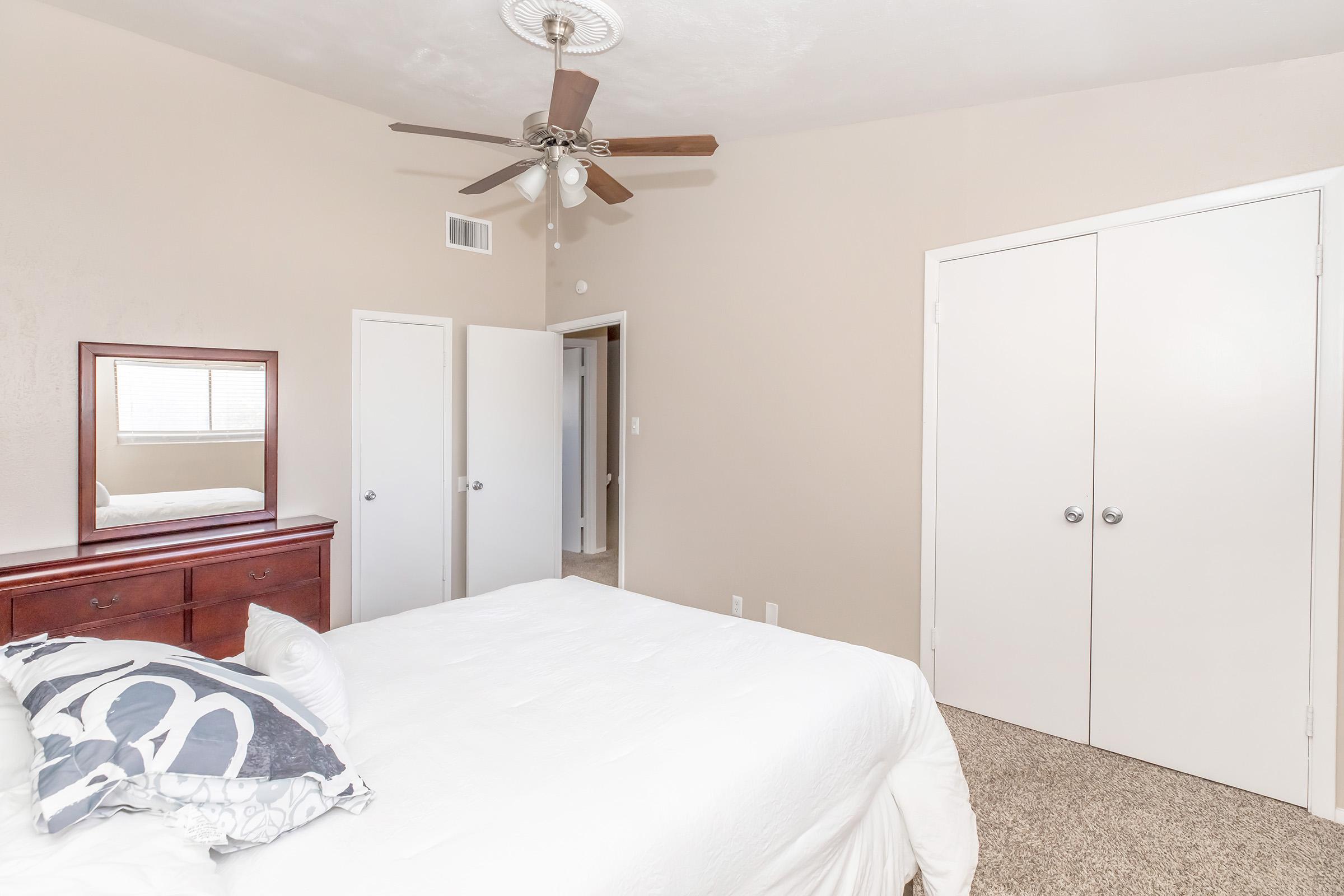 A cozy bedroom featuring a queen-sized bed with white bedding and decorative pillows. A wooden dresser with a mirror is positioned nearby. The room has a ceiling fan, beige walls, and light carpet. Two closed closet doors and an open door lead to another space, creating a spacious and inviting atmosphere.