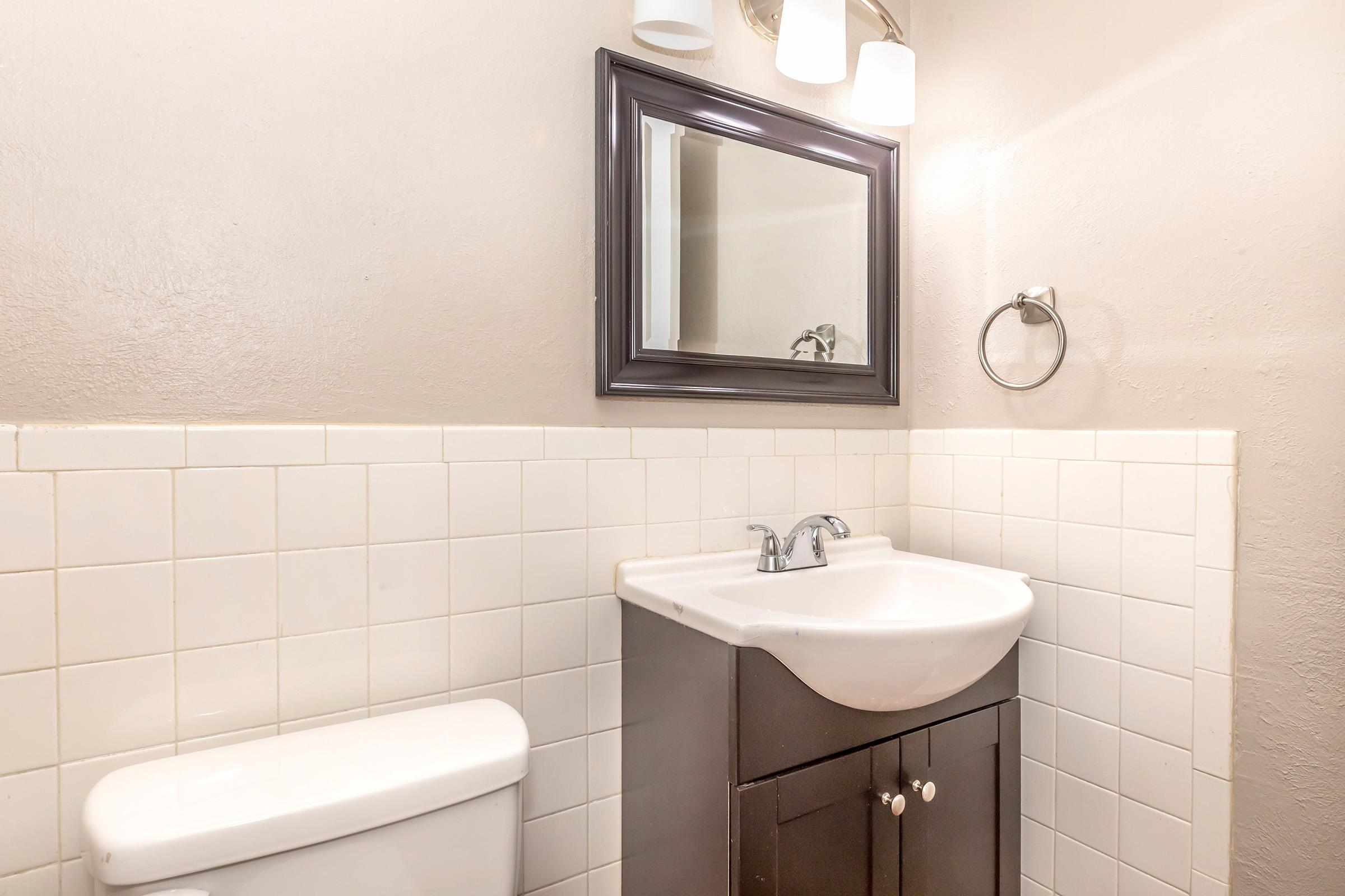 A small bathroom featuring a white sink with a dark cabinet underneath, a toilet beside it, and a large mirror above the sink. The walls are painted in a neutral tone, and there are light fixtures above the mirror. The tile backsplash consists of white square tiles.