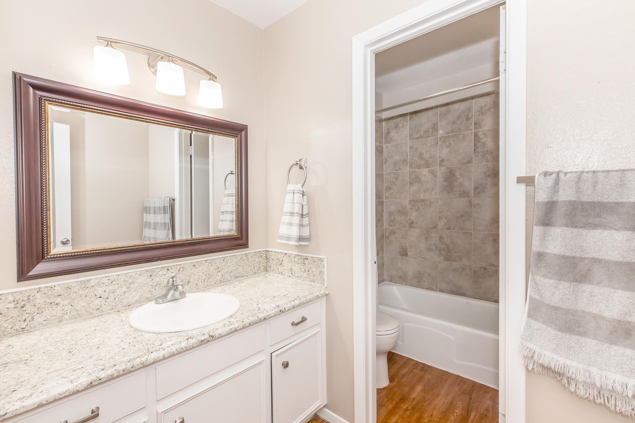 Modern bathroom featuring a granite countertop sink with a mirror above. Soft neutral walls and a tiled shower area can be seen in the background. A towel rack holds a striped towel, and the floor has a wood-like appearance, enhancing the warm atmosphere of the space.