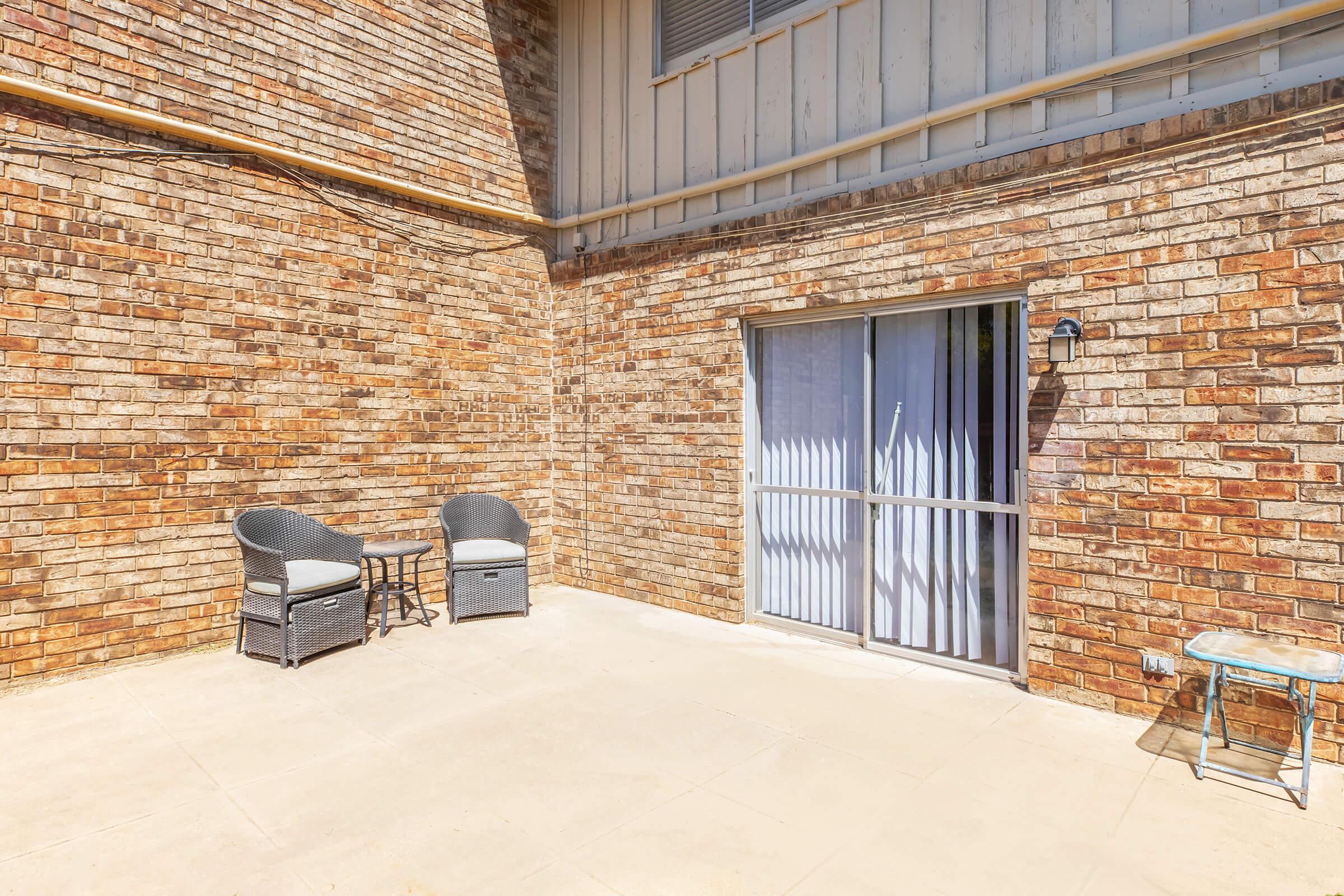 A spacious outdoor patio area featuring a brick wall, two wicker chairs with cushions, a small table, and a sliding glass door leading to the interior. Natural light illuminates the space, creating a warm and inviting atmosphere.