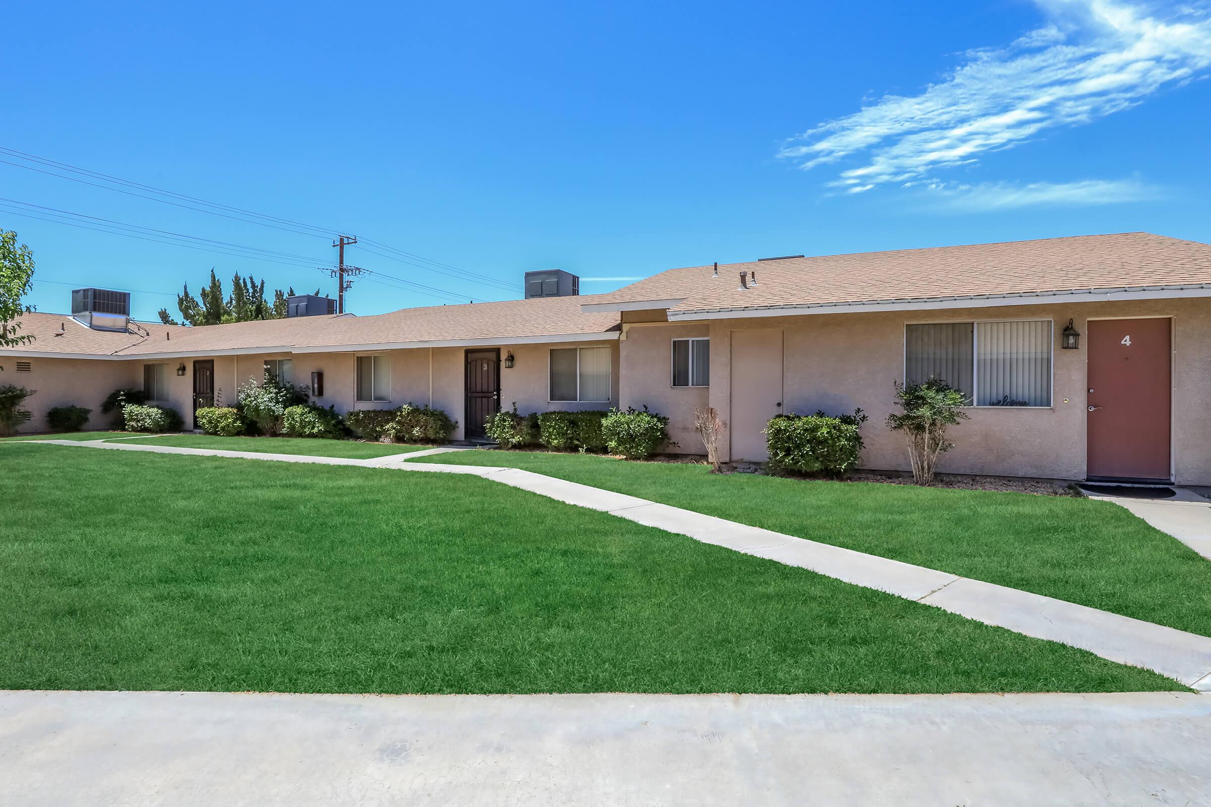 A single-story apartment complex with several units, featuring a well-maintained green lawn and concrete walkways. The building has a light-colored exterior and offers multiple doors, indicating different apartment entries, under a clear blue sky with some clouds.