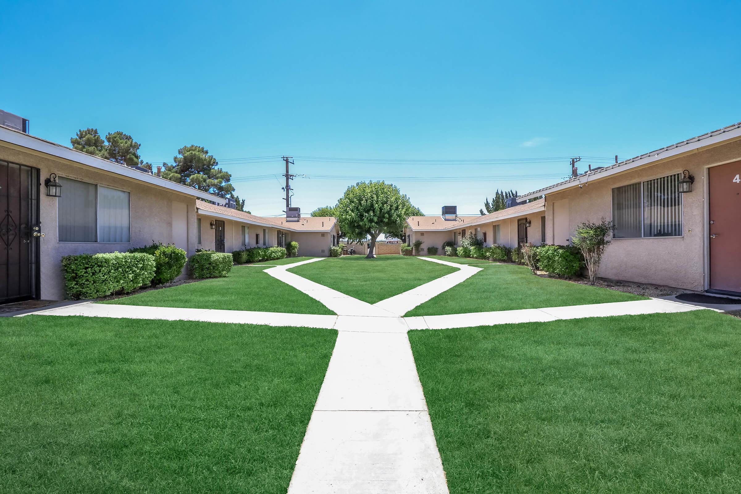 A sunny courtyard with well-maintained green grass and symmetrical pathways leading to doors of residential units. Small shrubs line the pathways, and trees provide shade, creating a tranquil atmosphere in a neighborhood setting. Clear blue sky overhead enhances the overall bright and inviting ambiance.