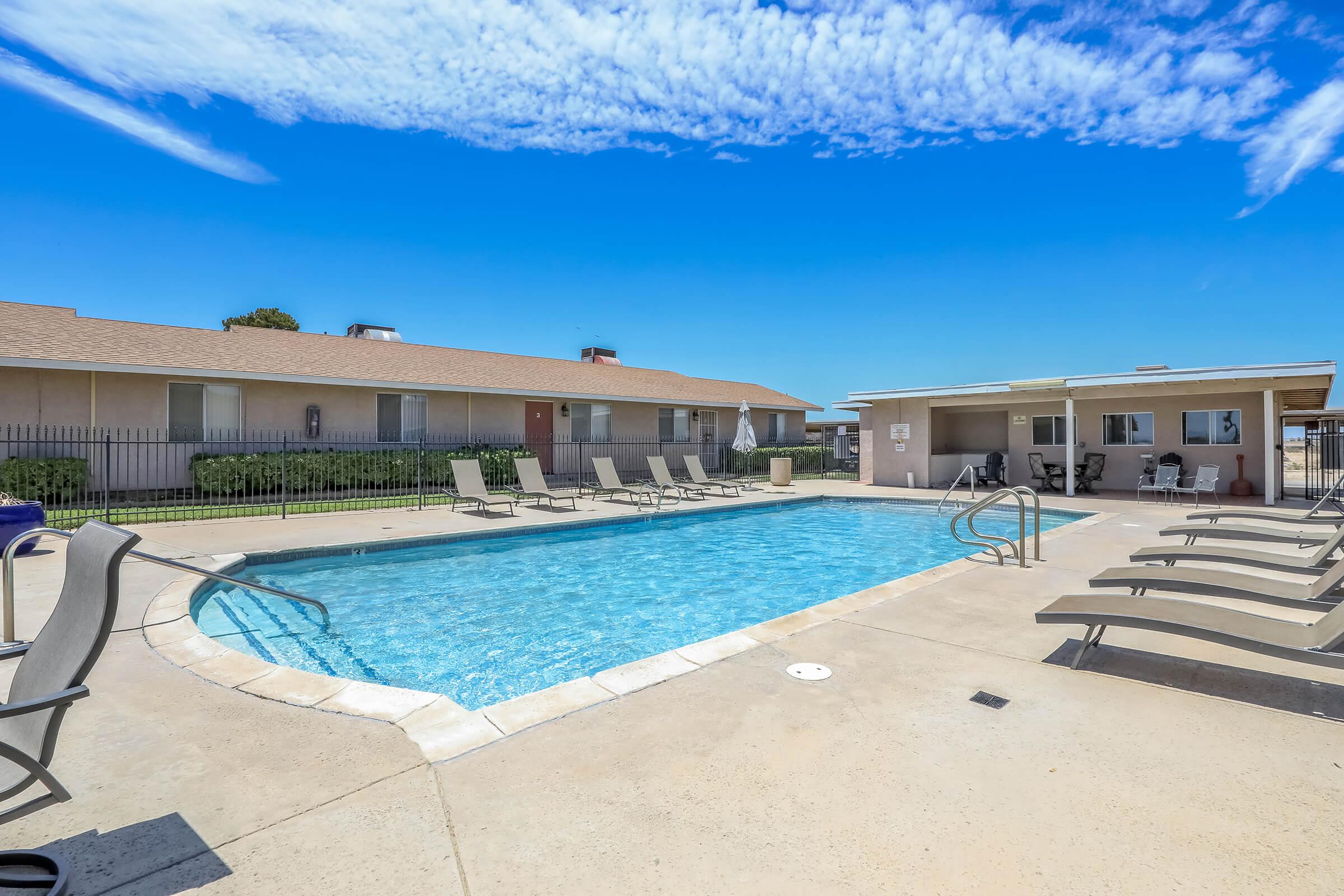 A clear blue swimming pool surrounded by lounge chairs and a grassy area, with a few apartment-style buildings in the background under a bright blue sky with wispy clouds. The scene is tranquil, ideal for relaxation and leisure.